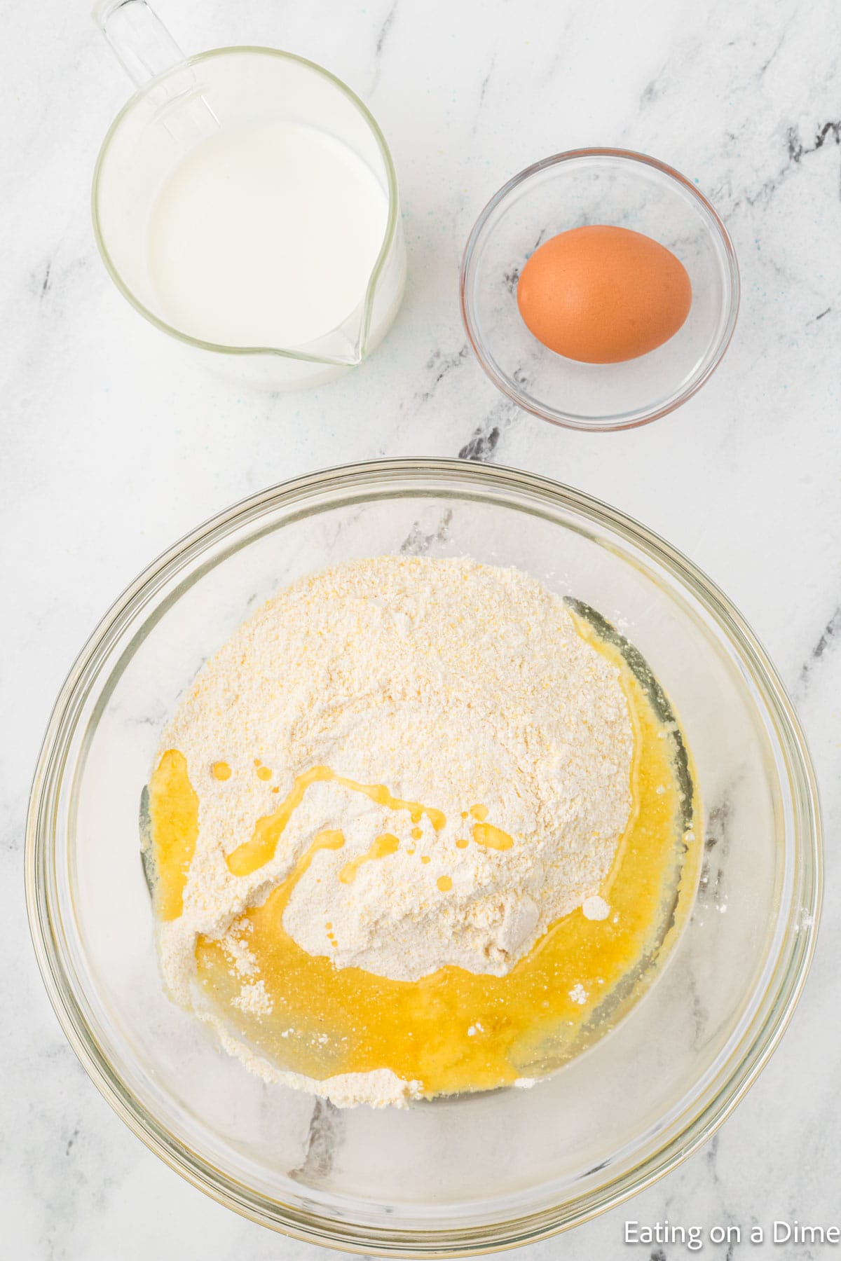 A glass bowl with flour and melted butter, a measuring cup of milk, and a single egg in a small bowl sit on the marble countertop, reminiscent of preparing ingredients for a comforting homemade hummus recipe.