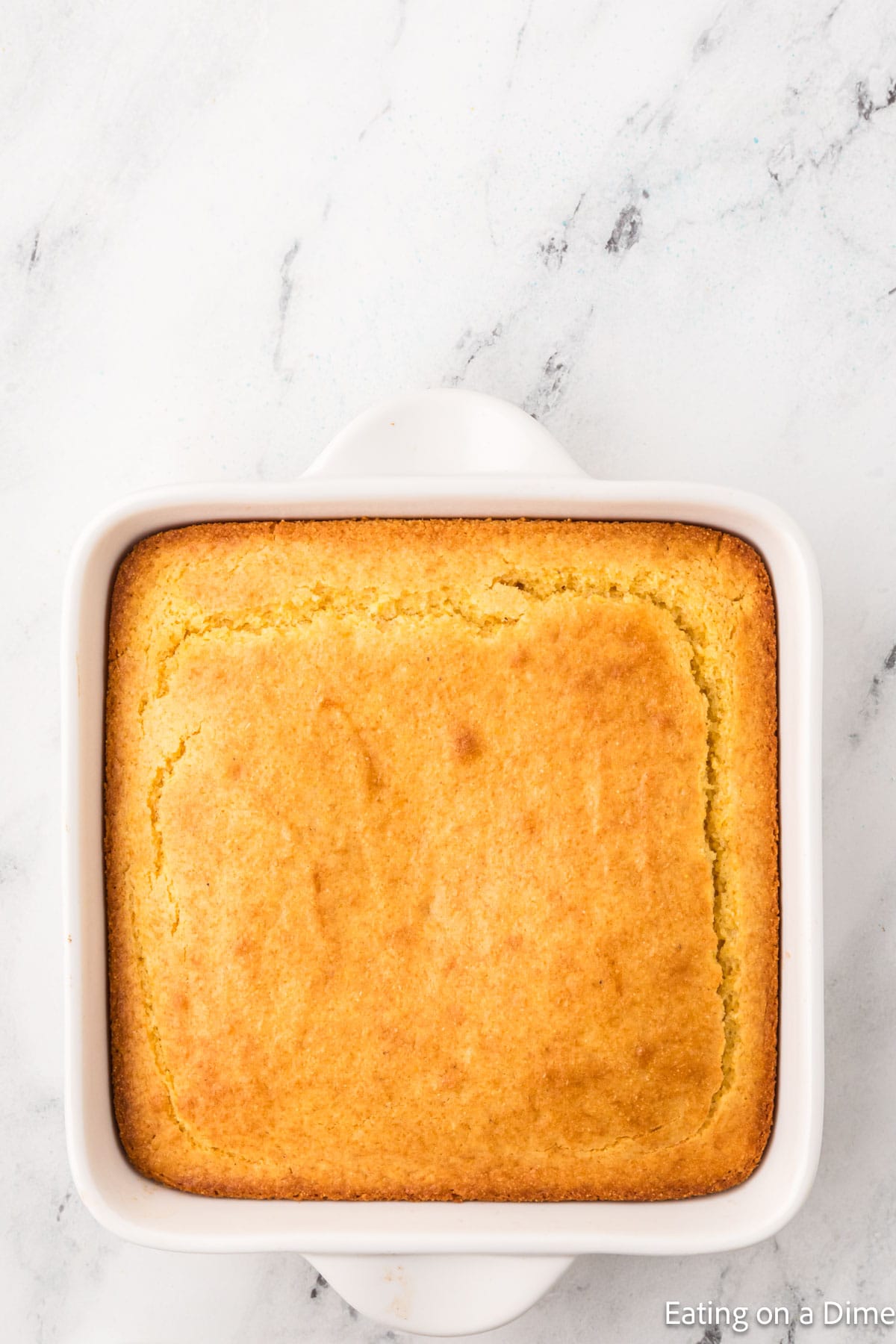 A square dish containing freshly baked, golden brown cornbread sits next to a bowl with a homemade hummus recipe. The cornbread is evenly cooked with a smooth top, resting on a white marble surface.