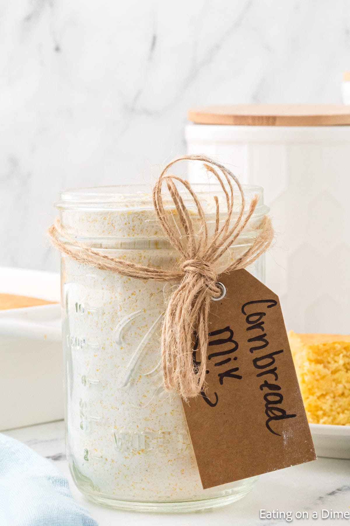 A mason jar filled with cornbread mix, tied with twine and a handwritten brown tag labeled "cornbread mix." The jar sits on a light-colored surface, accompanied by cornbread and a white dish partially visible in the background, offering a rustic touch reminiscent of a homemade hummus recipe.