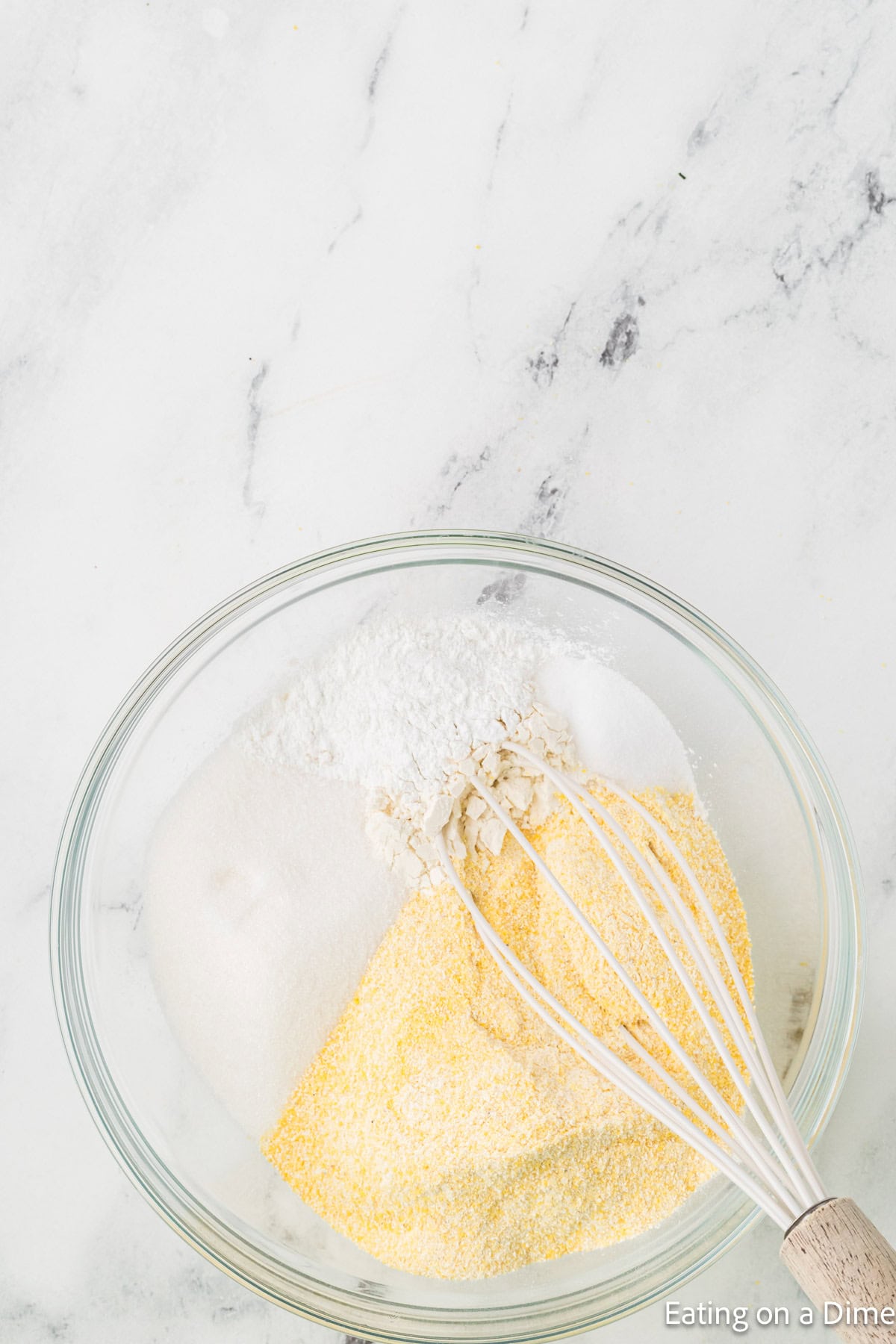 A glass bowl on a marble surface contains dry ingredients for cornbread: flour, cornmeal, sugar, baking powder, and salt. A whisk is partially visible in the bowl, poised to blend like a homemade hummus recipe awaiting its final touch.