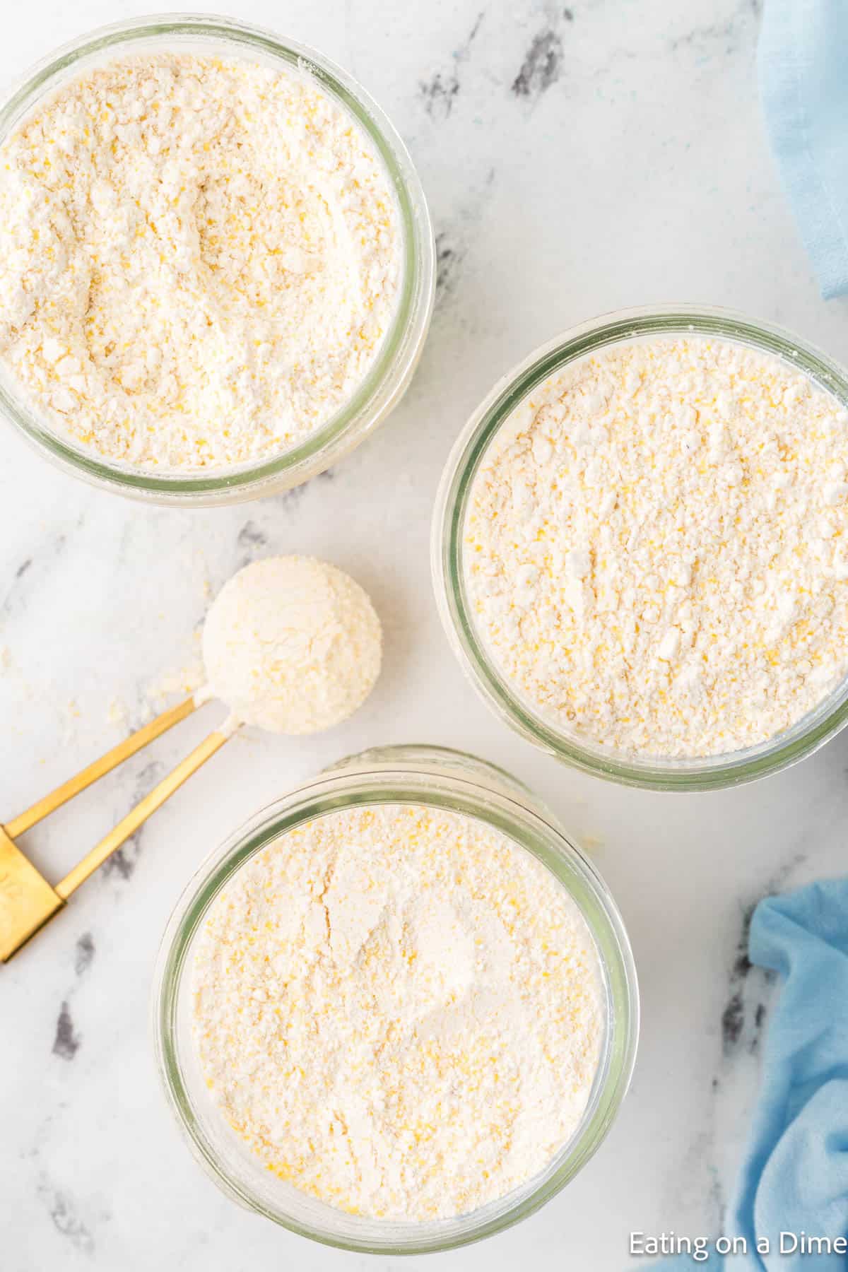 Three jars filled with a pale yellow, coarse powder for your homemade hummus recipe are arranged on a white marble surface. A gold measuring spoon, partially filled with the same powder, rests next to the jars. A light blue cloth is visible in the corner.