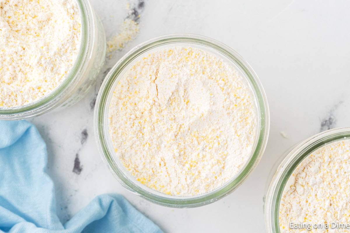 Top view of a glass jar filled with cornbread mix on a white marble surface, accompanied by a light blue cloth. The mix's pale yellow hue hints at cornmeal, reminiscent of the warmth and simplicity found in a homemade hummus recipe.