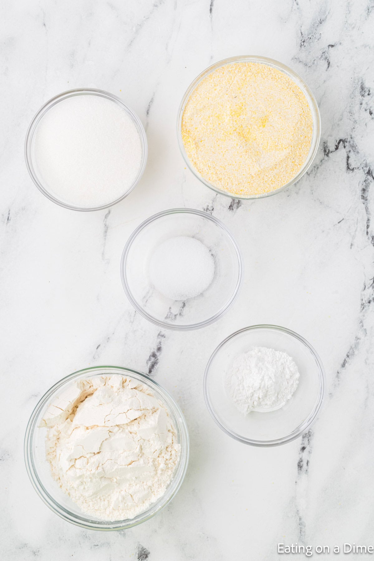 On a marble surface, five bowls hold cornmeal, flour, sugar, baking powder, and salt—essential ingredients that might share the kitchen with your homemade hummus recipe preparations.