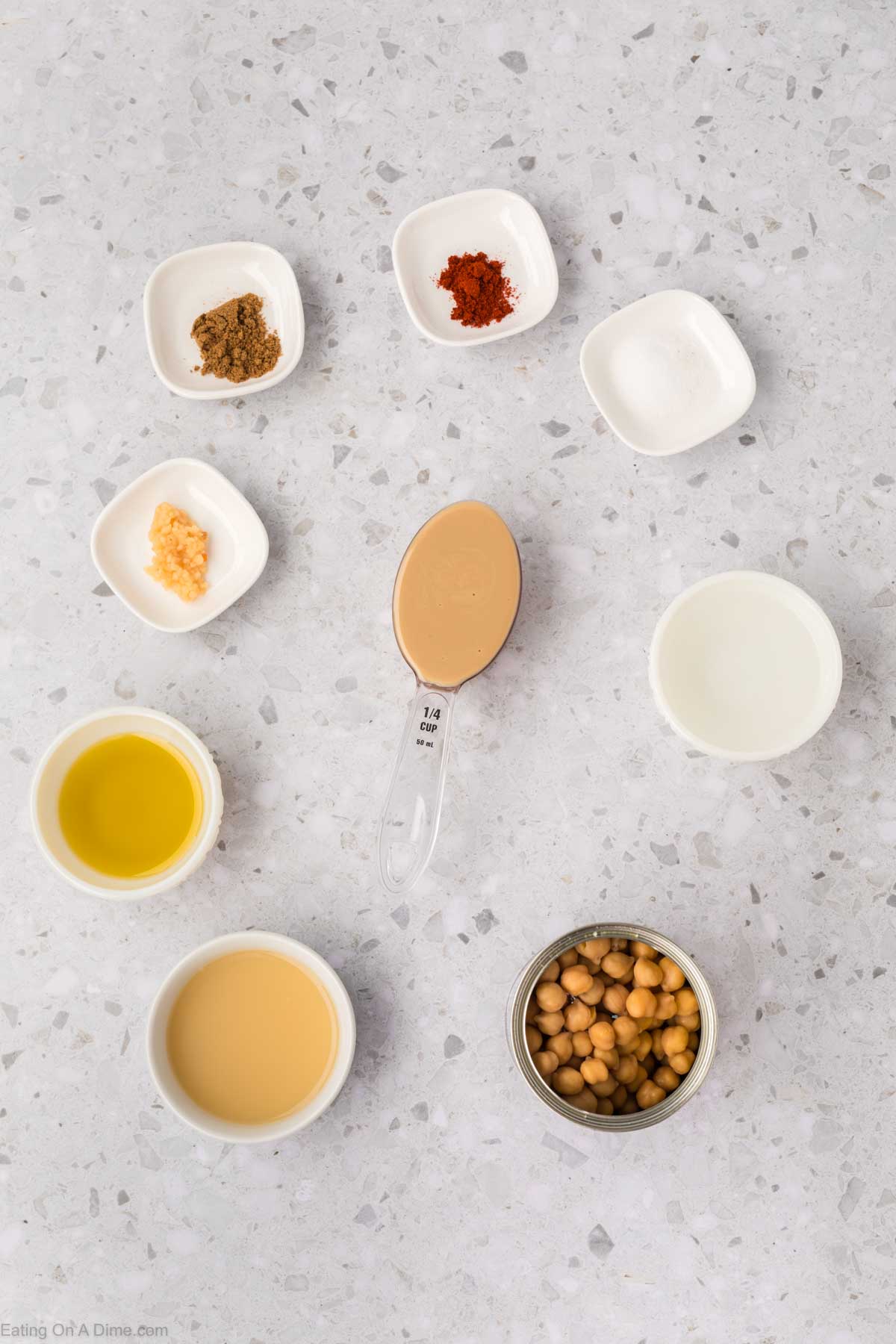 A flat lay of ingredients on a speckled countertop reveals the essentials for a homemade hummus recipe: spices in small bowls, a cup of chickpeas, a quarter-cup of tahini, minced garlic, oil, lemon juice, and water. Each is neatly arranged, ready to embark on your culinary adventure.