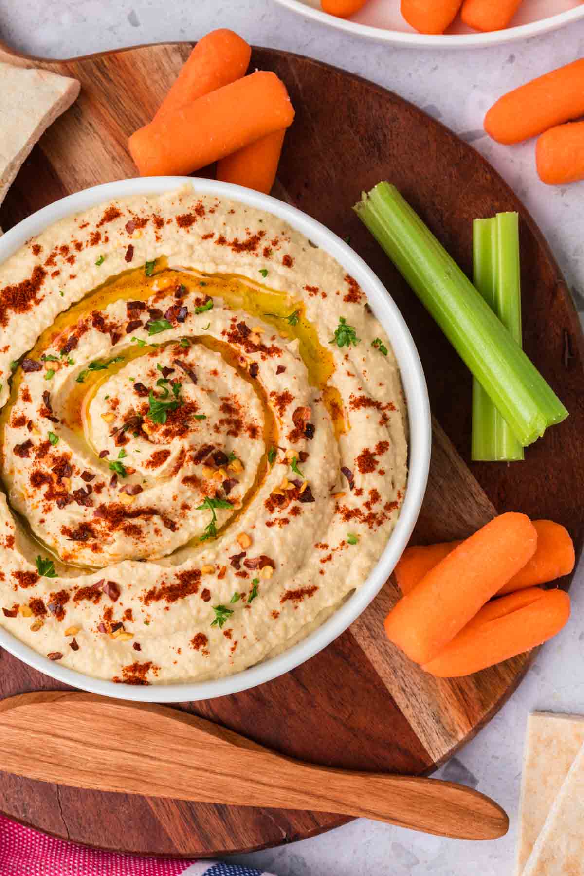 A bowl of creamy homemade hummus recipe is garnished with olive oil and paprika, served on a round wooden board. It is accompanied by baby carrots and celery sticks. A wooden spoon rests nearby, ready for serving.