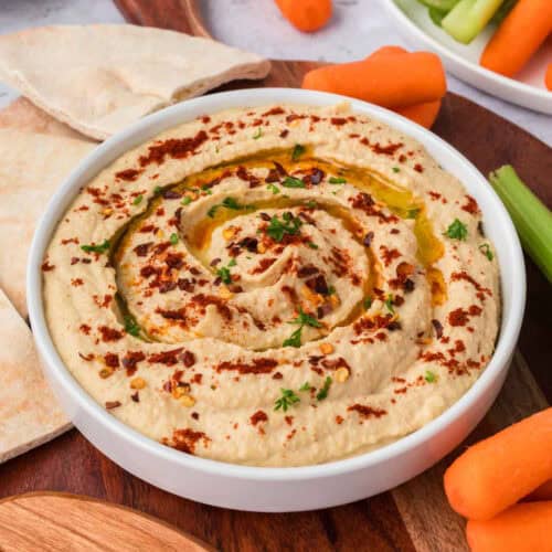 A bowl of creamy, homemade hummus recipe is garnished with paprika, olive oil, and herbs. It is surrounded by carrot sticks, celery, and pita bread pieces on a wooden board.