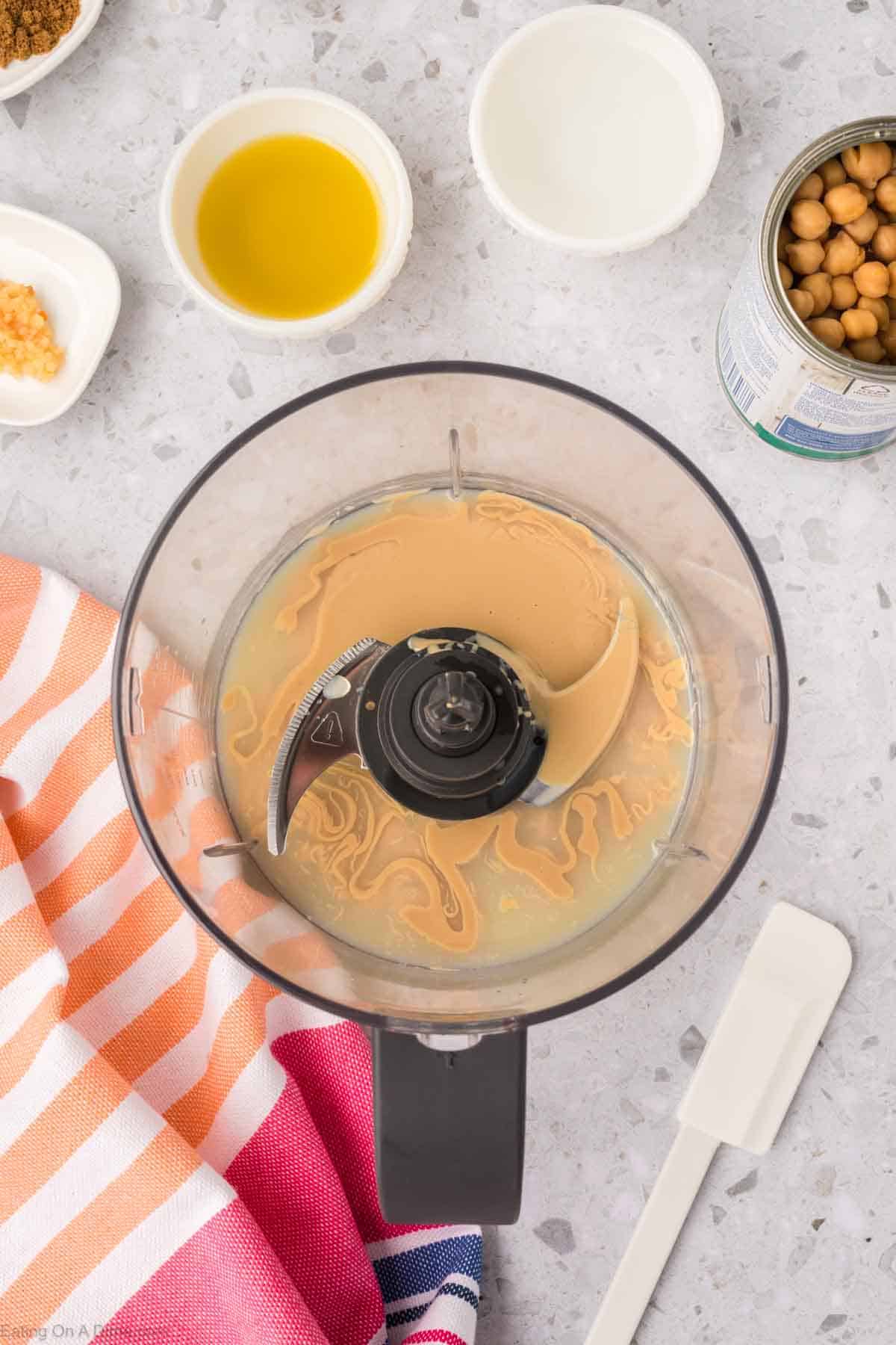 A food processor bowl with tahini inside, surrounded by bowls of olive oil, lemon juice, chickpeas, and minced garlic on a speckled countertop—a perfect setup for crafting your homemade hummus recipe. A striped cloth and a white spatula are placed next to the processor.