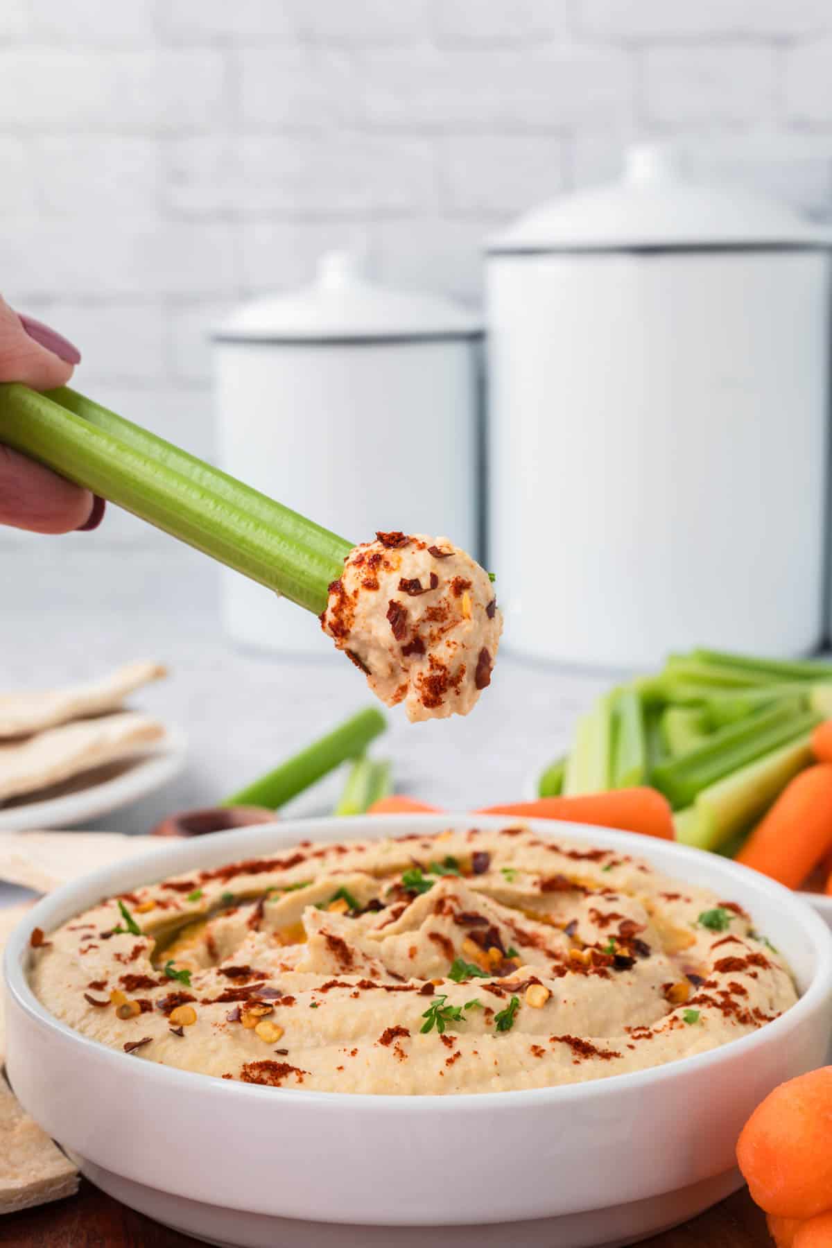 A hand dips a celery stick into a bowl of homemade hummus topped with spices and herbs. Carrot sticks and more celery are visible in the background, alongside white kitchen containers on the counter.