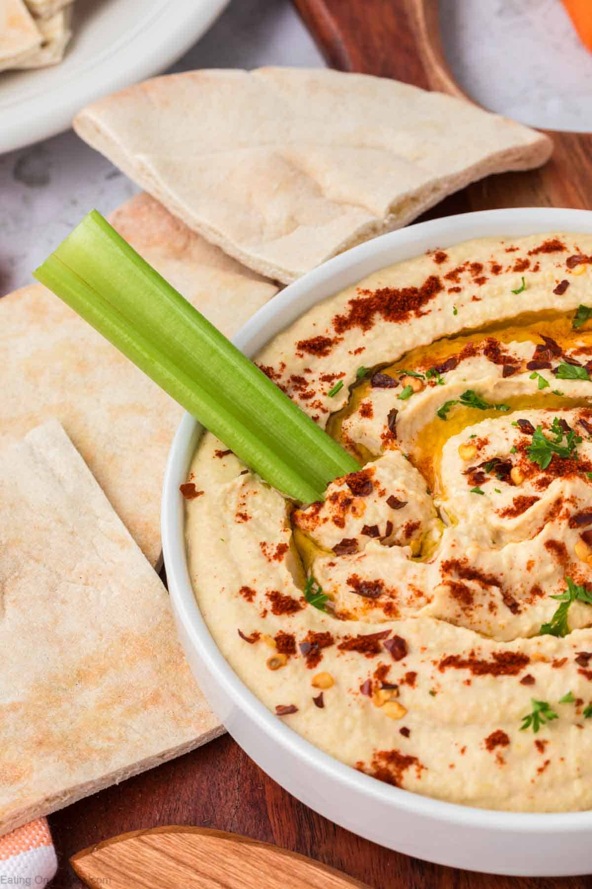 A white bowl filled with creamy homemade hummus, garnished with paprika and parsley, with a celery stick on top. Sliced pita bread is placed next to the bowl on a wooden board.