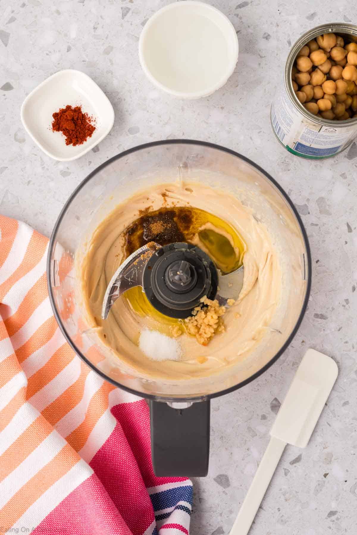 A top view showcases a food processor with ingredients for a homemade hummus recipe, including olive oil, garlic, and spices. Nearby lie a striped towel, spatula, can of chickpeas, small dish of paprika, and an empty bowl on a speckled countertop.