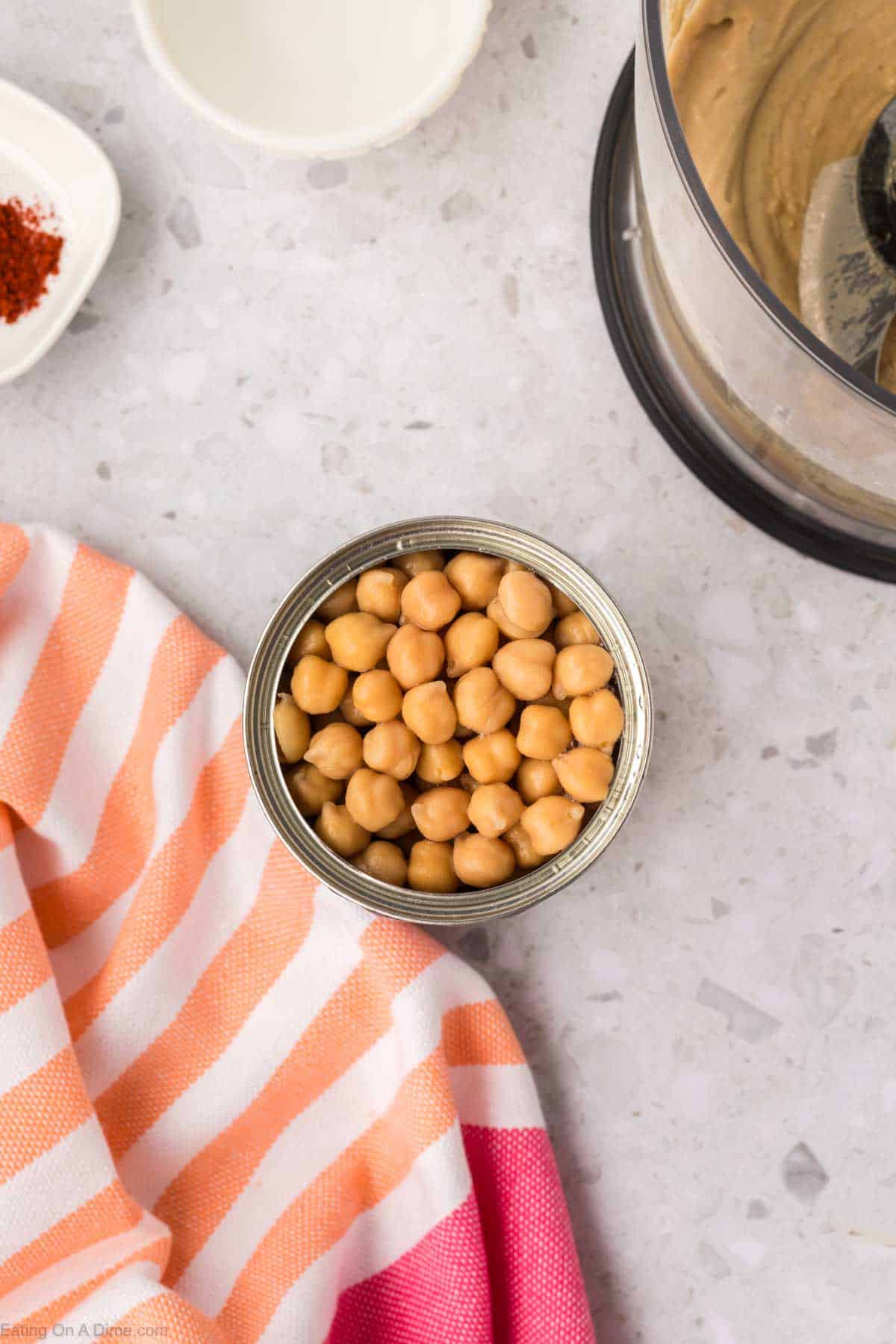 An open can of chickpeas sits on a light gray countertop beside a striped orange and pink cloth, with a food processor partially filled with creamy hummus, hinting at a delicious homemade hummus recipe. A small dish with red spice adds an extra touch in the background.