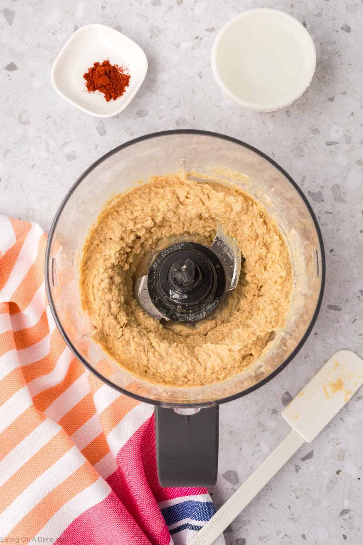 A food processor containing a creamy beige mixture of homemade hummus sits on a speckled countertop. Nearby are a small bowl of red spice, a white bowl, a rubber spatula, and a colorful striped cloth.