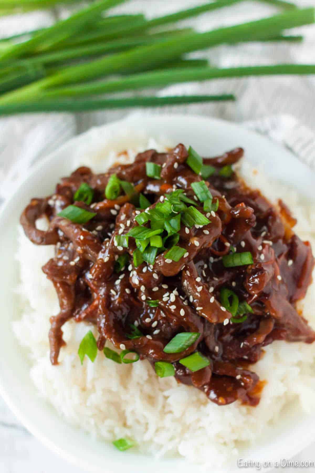 A dish of savory slow cooker Mongolian beef stir-fry topped with sesame seeds and chopped green onions is served over a mound of white rice. Fresh green onions are placed in the background.