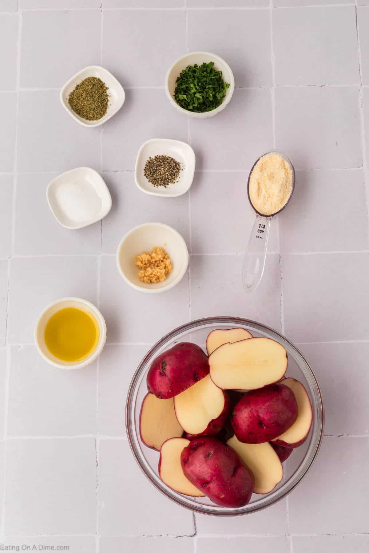 A bowl of sliced red potatoes on a tiled surface, ready to become delicious roasted red potatoes, is surrounded by small bowls and a spoon containing herbs, garlic, oil, seasoning, and grated cheese.