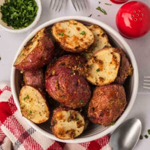 A bowl of perfectly roasted red potatoes, seasoned with garlic and herbs, garnished with chopped parsley. The bowl sits on a table adorned with a red and white napkin, silverware, a small bowl of herbs, and charming red salt and pepper shakers nearby.