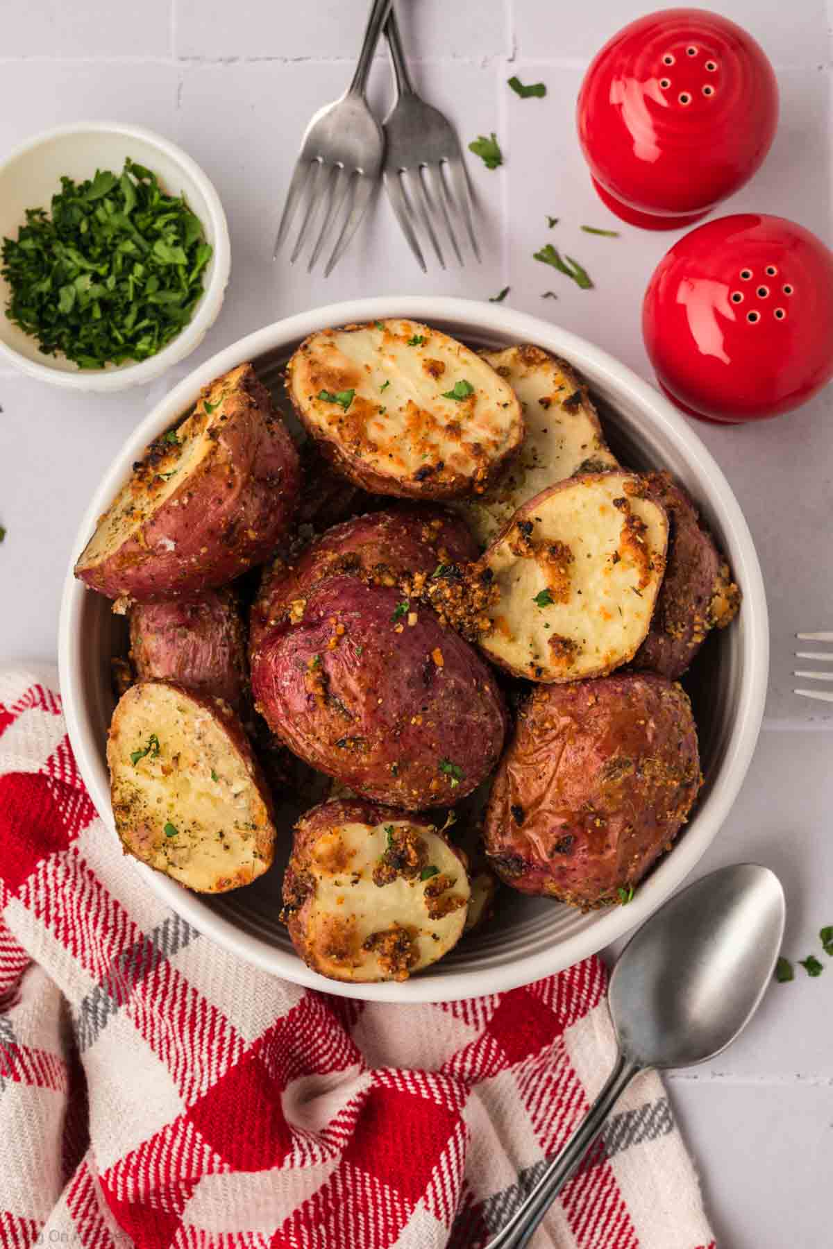 A bowl of roasted red potatoes seasoned with herbs sits on a table. Next to these savory delights are a small bowl of chopped parsley, two forks, a spoon, salt and pepper shakers, and a red and white checkered cloth.