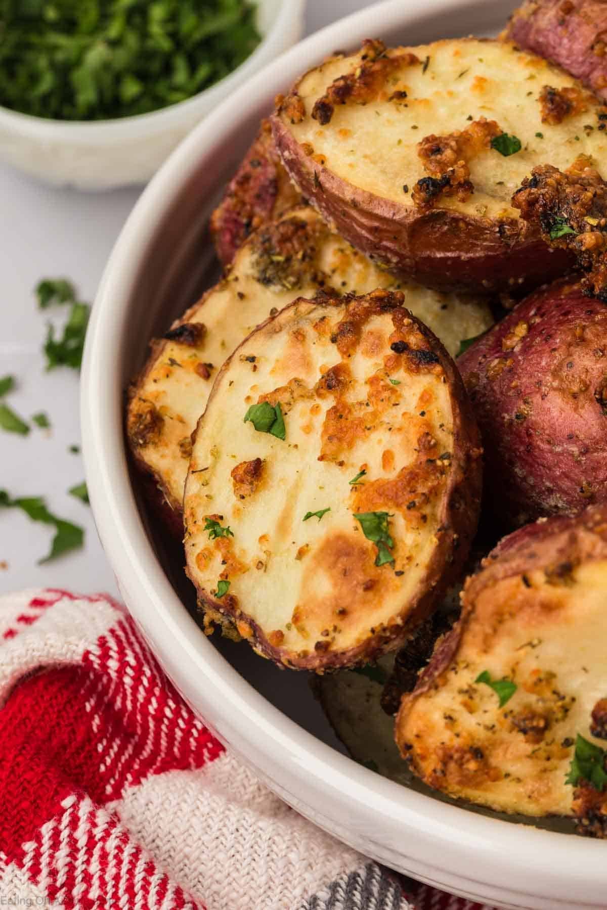 A bowl of roasted red potatoes halves seasoned with herbs and spices. The potatoes are golden brown with visible herbs on top. A red and white striped cloth is beside the bowl, and a small bowl of chopped parsley is partially visible in the background.