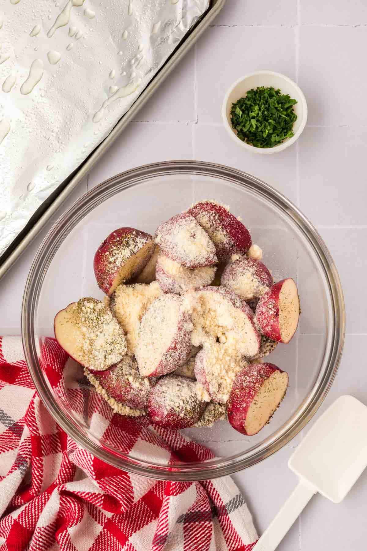 A glass bowl filled with halved red potatoes, soon to become delicious roasted red potatoes, is coated in a seasoning mixture. A small bowl of chopped herbs is nearby. A red and white checkered cloth is next to the bowl, and a baking sheet waits eagerly in the background.