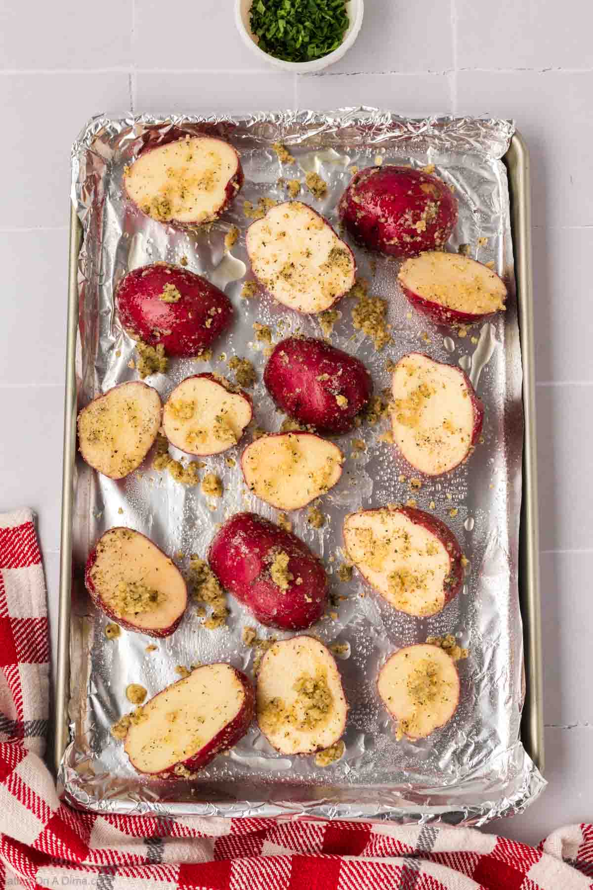 Roasted red potatoes, halved and seasoned with breadcrumbs, rest on a foil-lined baking tray. A small bowl of chopped herbs is in the background, accentuating the aroma, while a red and white checkered cloth peeks from the corner.