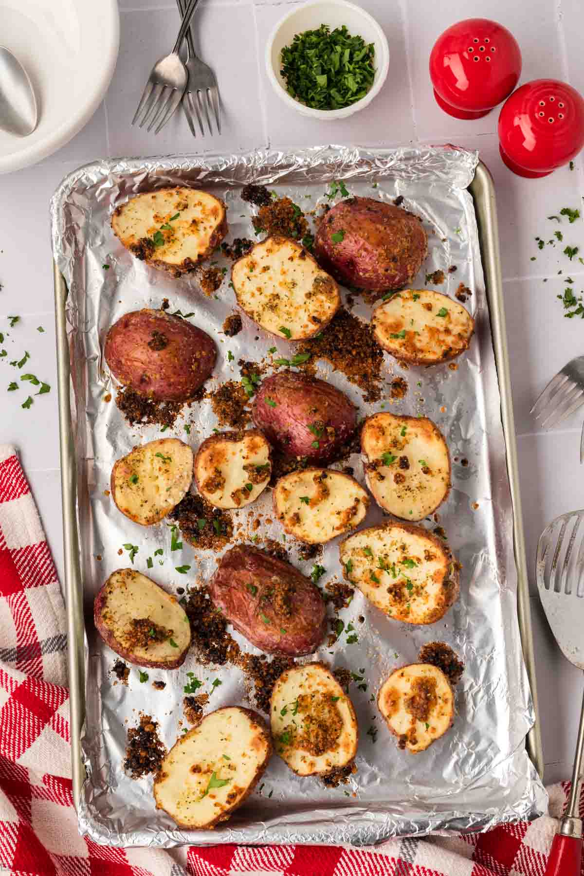 A baking sheet lined with foil holds several roasted red potato halves, sprinkled with herbs and breadcrumbs. Nearby are a red and white checkered cloth, a bowl of chopped herbs, red salt and pepper shakers, and utensils.