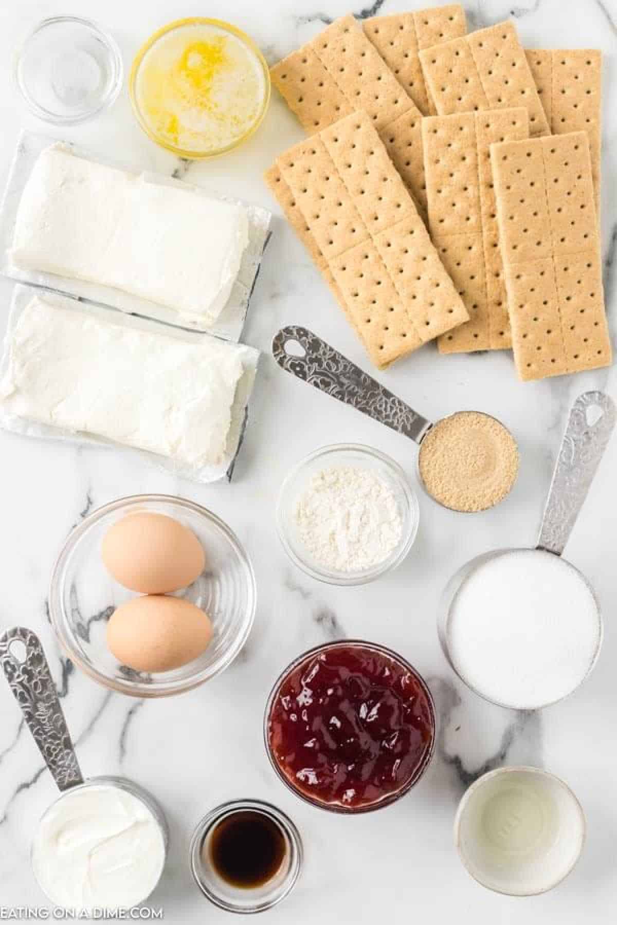 A flat lay of ingredients on a marble surface, including cream cheese blocks, graham crackers, eggs, butter, sour cream, vanilla extract, sugar, flour, brown sugar, and a jar of strawberry jam—perfect for whipping up raspberry cheesecake bars. Measuring cups and spoons are also present.