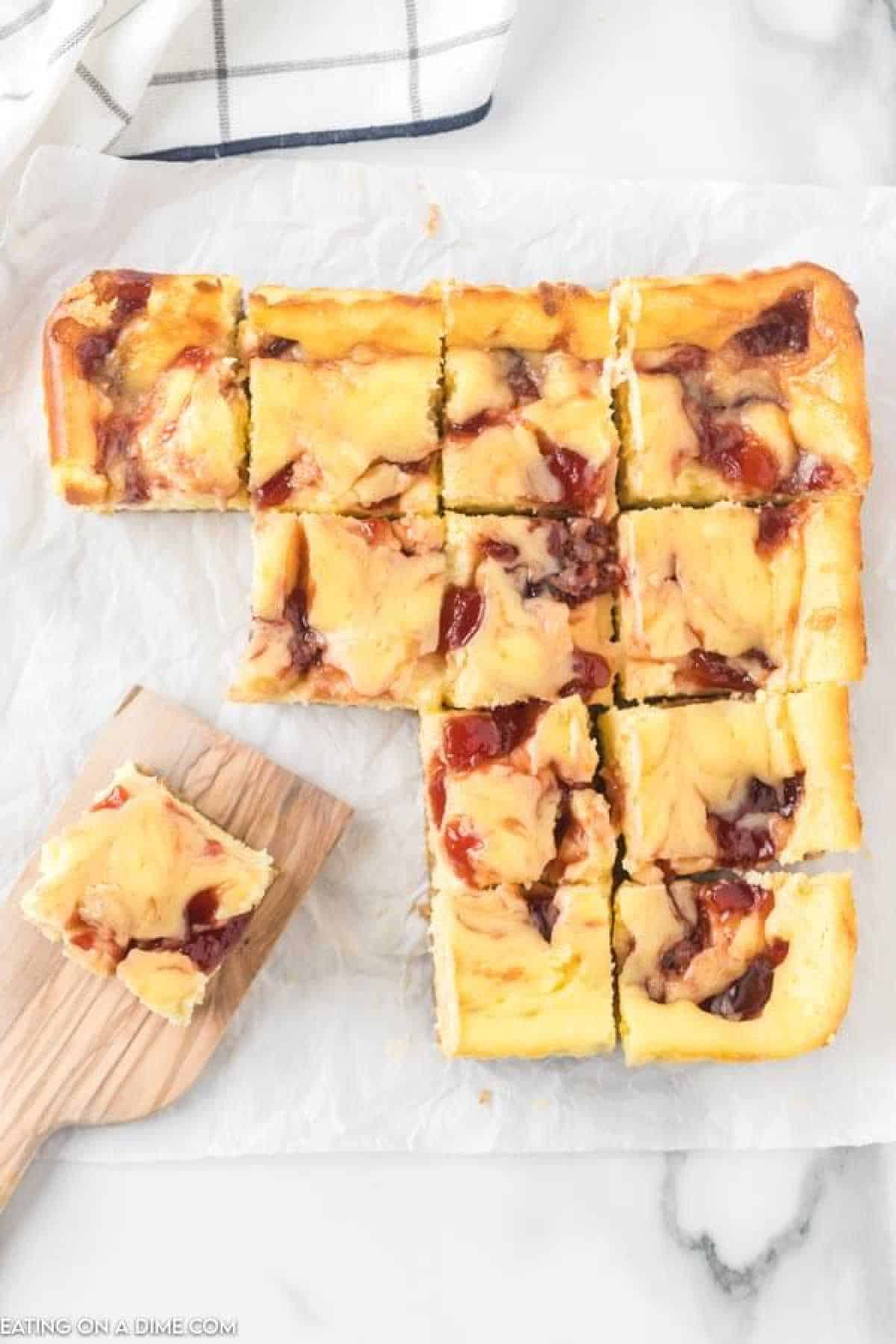 A batch of raspberry cheesecake bars with a cherry swirl, cut into 12 square pieces, is displayed on a piece of parchment paper. One bar is placed on a wooden spatula, ready to be served. A white and black checkered cloth is partially visible in the background.