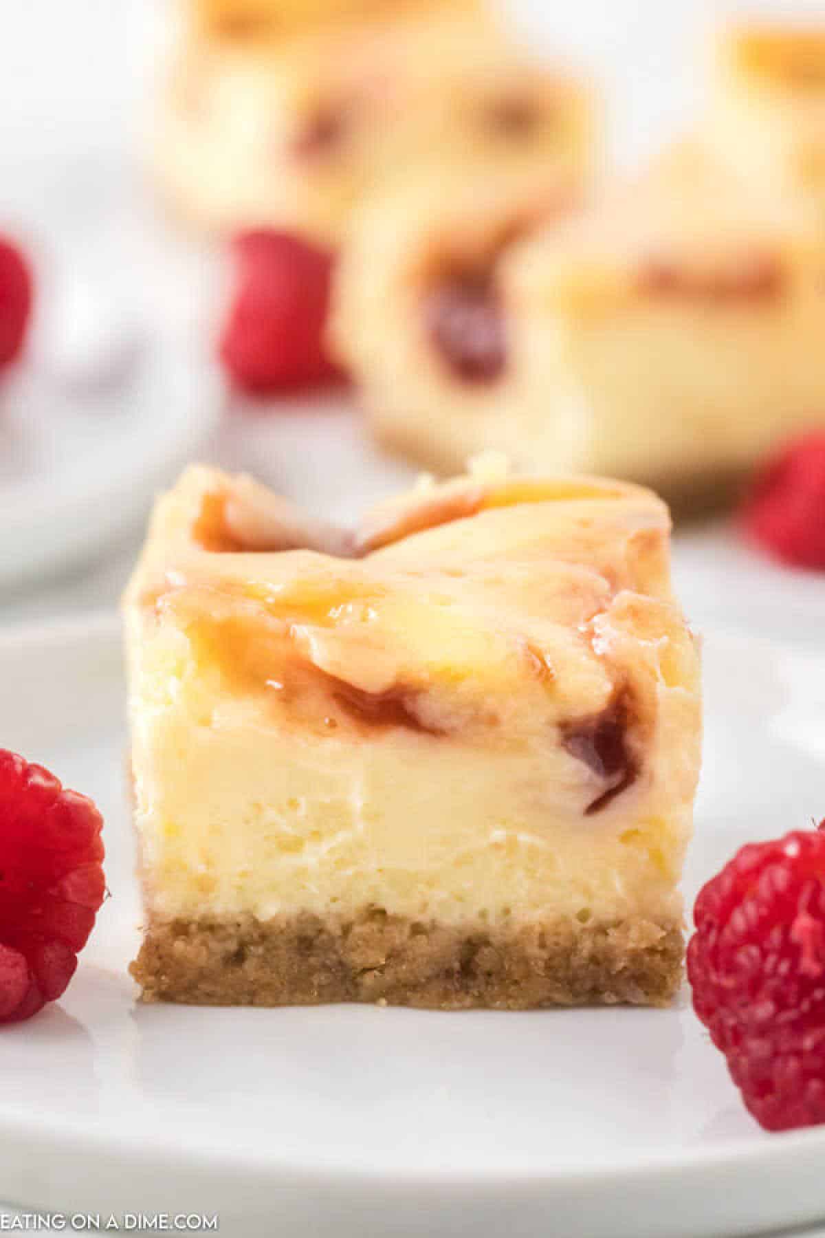 A close-up of a slice of creamy raspberry cheesecake bar with a swirl of raspberry on top. The dessert has a graham cracker crust and is garnished with fresh raspberries on a white plate. Other slices of the raspberry cheesecake bars and additional raspberries are blurred in the background.