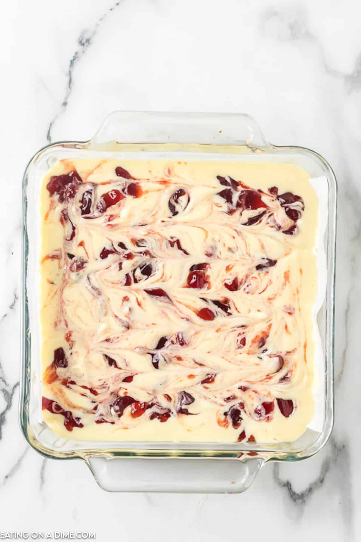 A square, glass baking dish filled with a creamy white mixture that has ripples of red, likely a swirl of jam or fruit preserves, spread throughout. These raspberry cheesecake bars sit invitingly on a white marble surface.