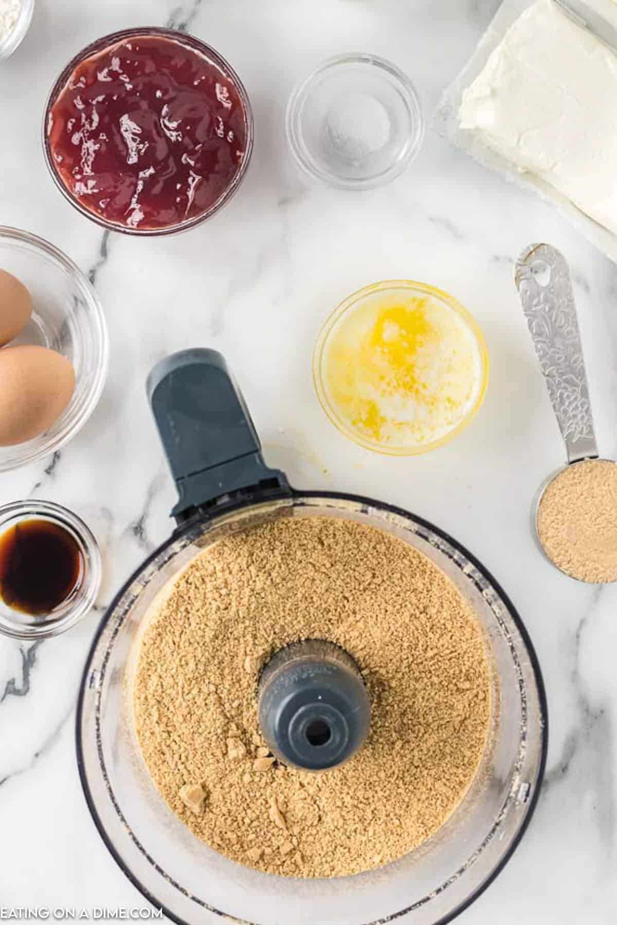 Top-down view of ingredients for raspberry cheesecake bars. A food processor with crushed graham crackers, a bowl of melted butter, a small measuring spoon with sugar, vanilla extract, two eggs, a bowl of strawberry sauce, and a block of cream cheese are arranged on a countertop.