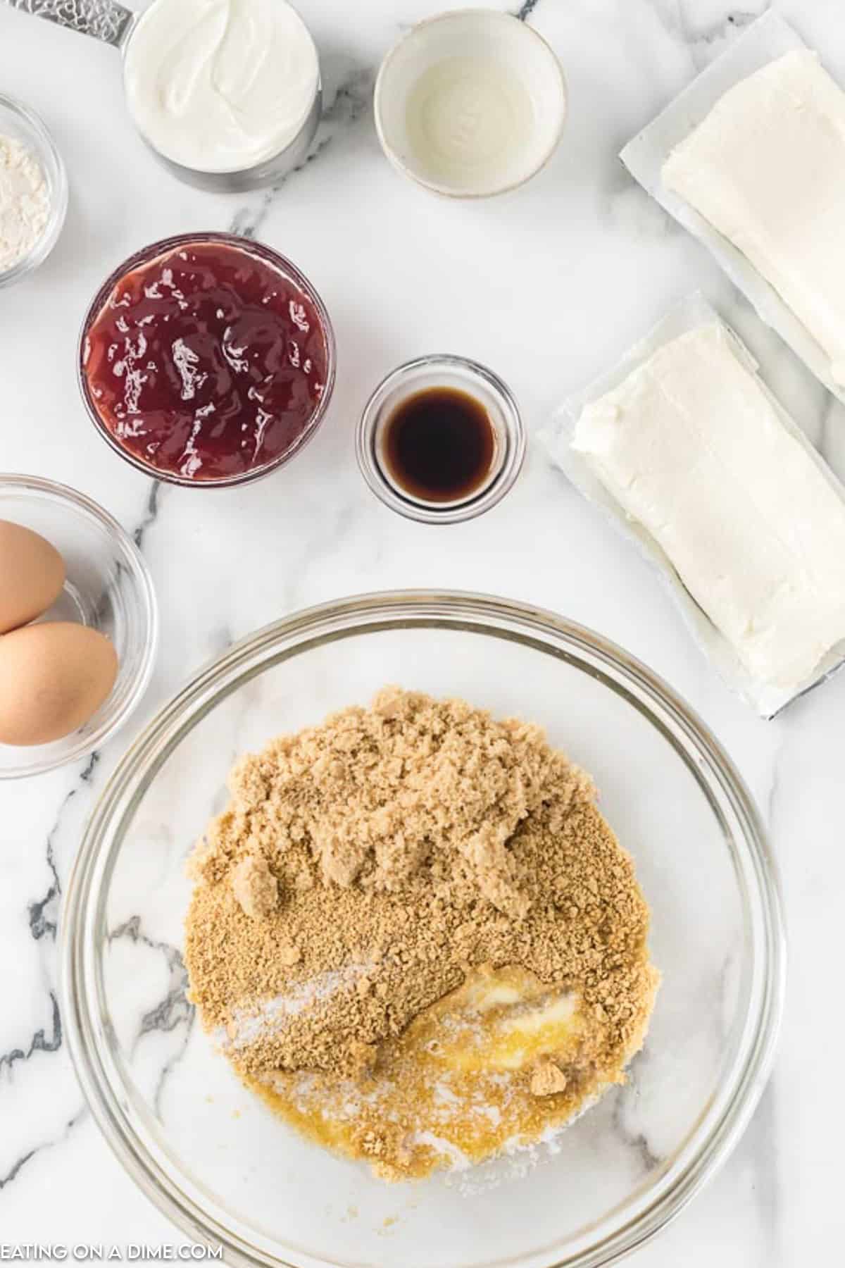 A top-down view of ingredients for raspberry cheesecake bars on a marble countertop. Items include a bowl with brown sugar and melted butter, blocks of cream cheese, a glass bowl of cherry pie filling, two eggs, a small bowl of vanilla extract, and a container of sour cream.