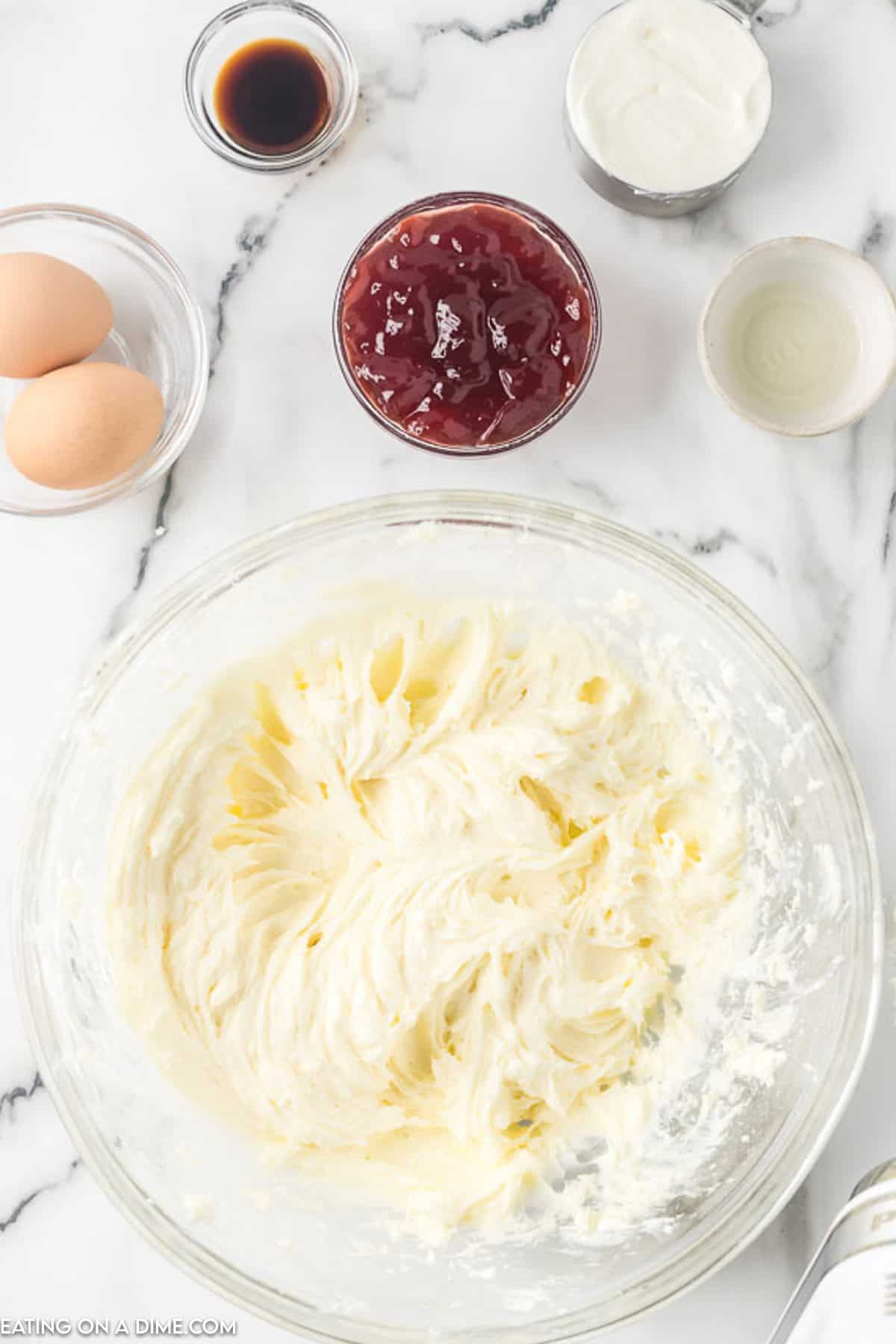 A glass mixing bowl filled with creamy white batter, destined to become delicious raspberry cheesecake bars, sits on a marble countertop. Surrounding it are two eggs, a small bowl of dark liquid, a bowl of white cream, a jar of red jam, and an empty small bowl. A silver spoon is partially visible in the batter.