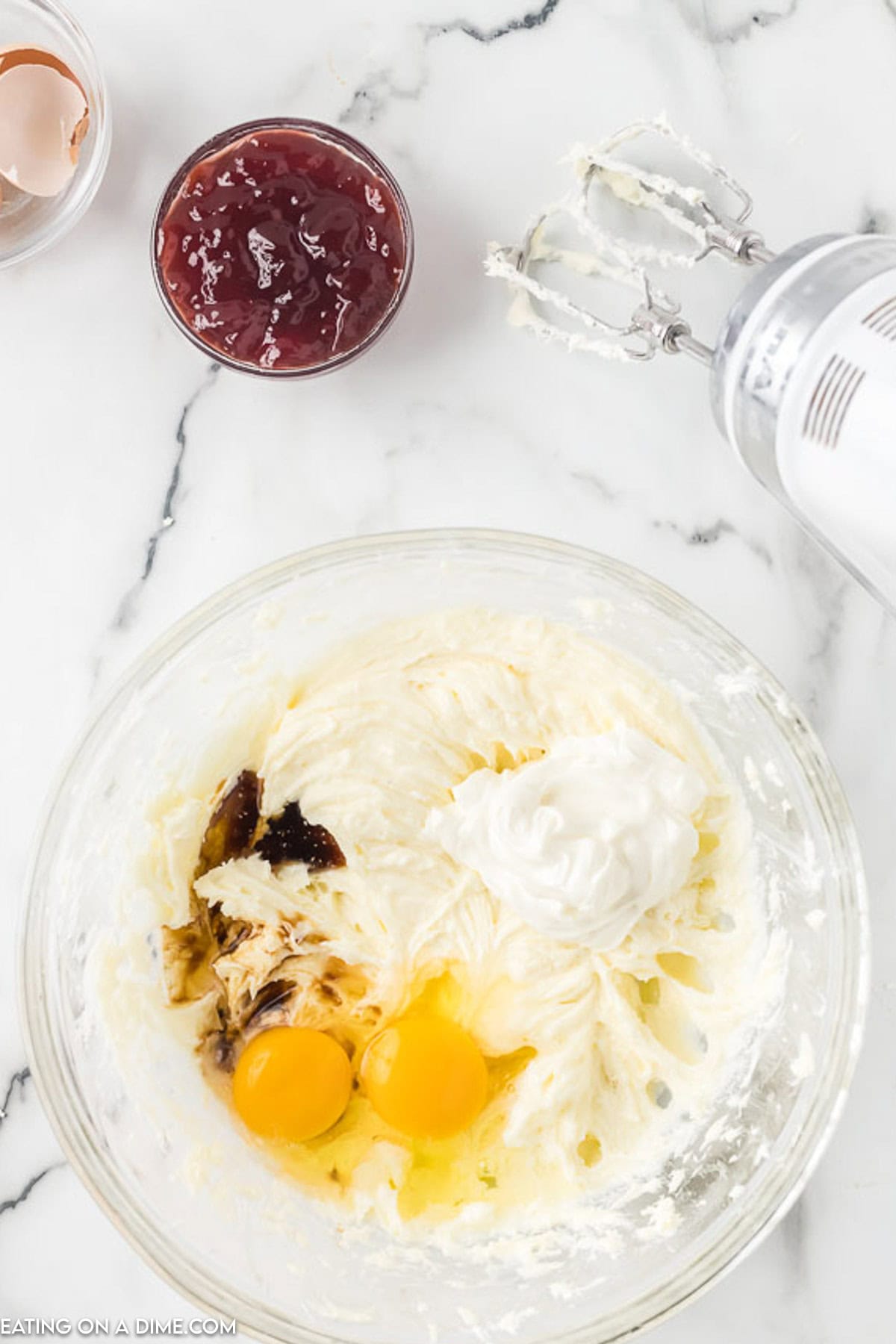 A clear glass mixing bowl on a marble countertop contains a creamy mixture with two whole eggs and a dollop of sour cream, soon to be the base for raspberry cheesecake bars. An electric mixer with beaters is partially visible next to the bowl. A small bowl of jam and a cracked egg shell are nearby.