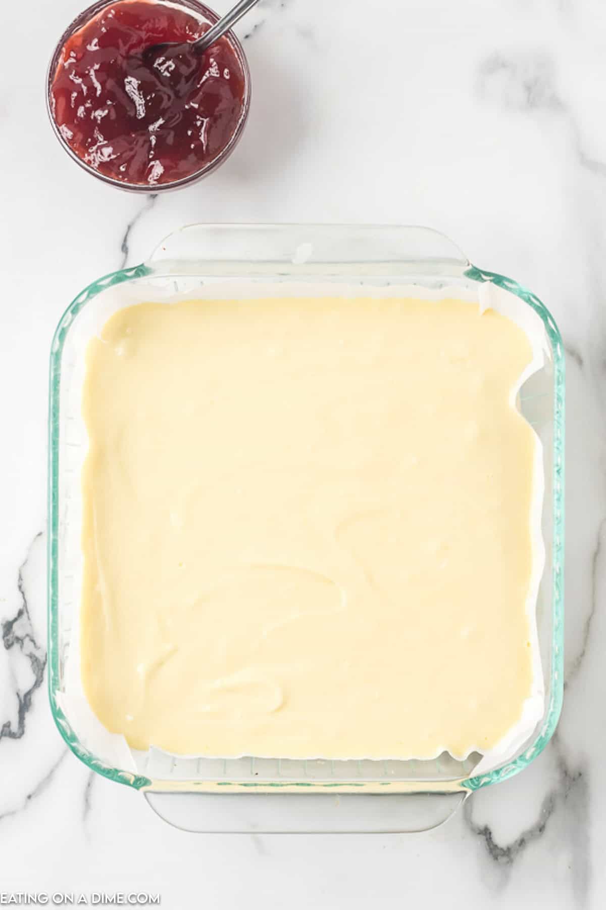 A square glass baking dish filled with a layer of smooth batter sits on a white marble surface. Above the dish is a small glass bowl containing red jam, perfect for topping your homemade raspberry cheesecake bars.