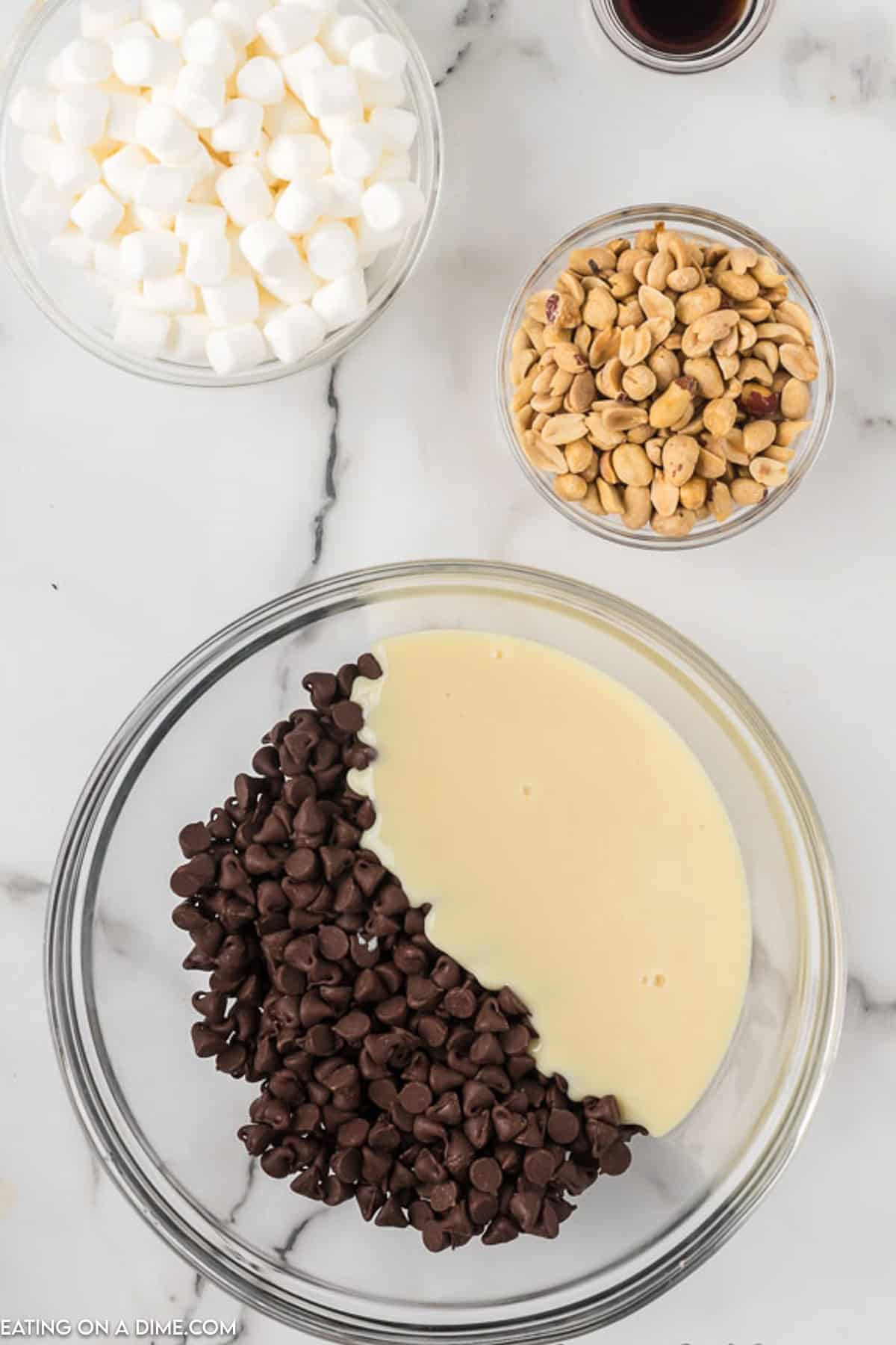 A top view of ingredients for an easy rocky road fudge recipe on a marble countertop. There's a large bowl with chocolate chips and condensed milk, a smaller bowl with marshmallows, another small bowl of peanuts, and a tiny bowl of vanilla extract. The items are neatly arranged.