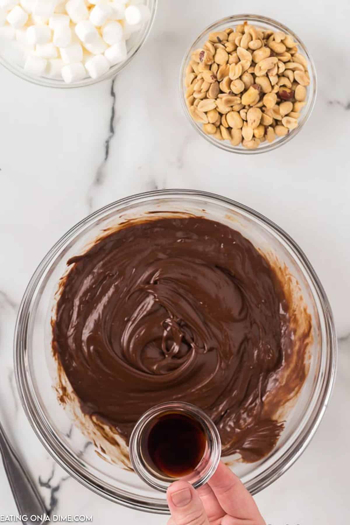 A glass bowl filled with melted chocolate sits on a marble countertop. Surrounding it are a small bowl of vanilla extract being held, as well as bowls of mini marshmallows and peanuts—essential ingredients for an easy rocky road fudge recipe. A black utensil rests next to the chocolate bowl.