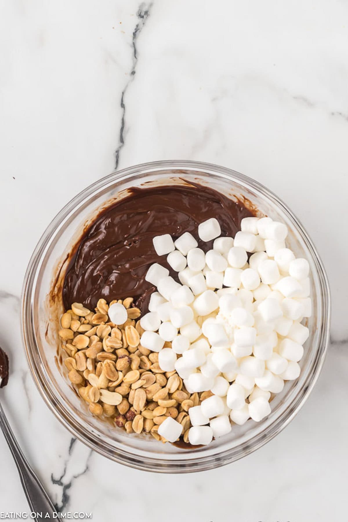 A clear glass bowl on a marble countertop contains a partially mixed combination of melted chocolate, mini marshmallows, and peanuts—perfect for an easy rocky road fudge recipe. A spoon rests beside the bowl.