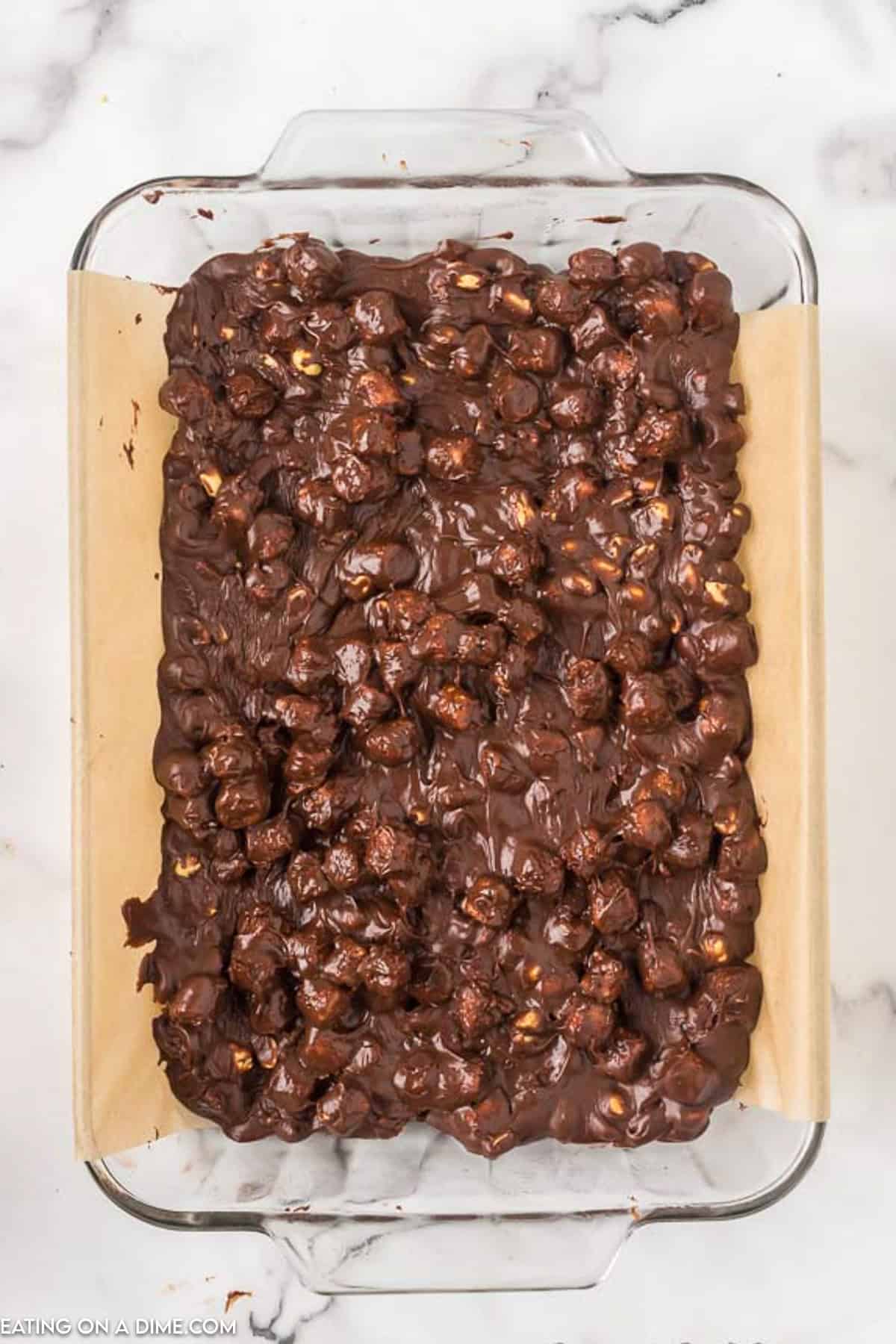 A glass baking dish filled with a mixture of melted chocolate, marshmallows, and nuts on top of parchment paper. This easy rocky road fudge recipe features rich, gooey chocolate with chunks of marshmallows and bits of nuts beautifully embedded throughout.