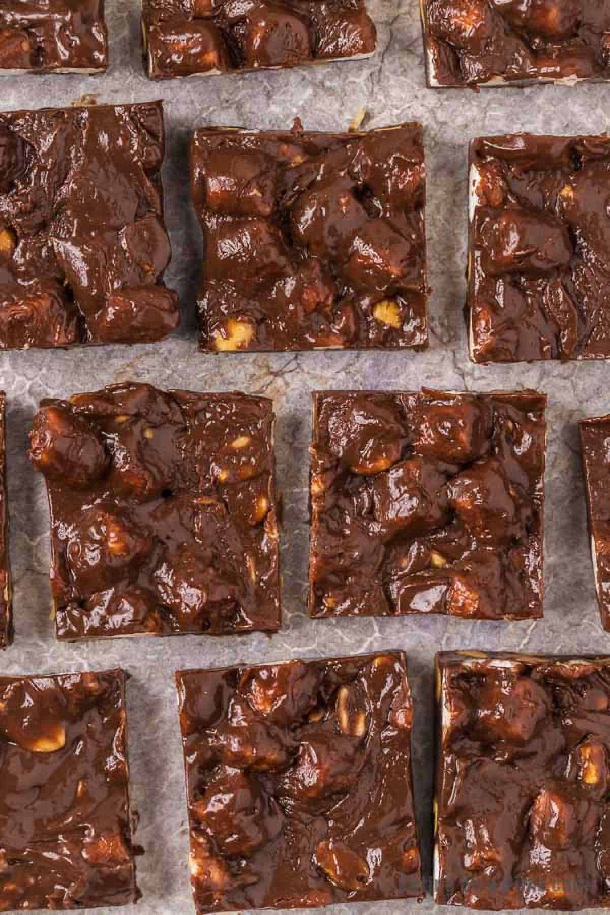 A close-up image of multiple square pieces of homemade rocky road fudge with nuts, arranged neatly on parchment paper. The fudge has a rich, glossy texture with visible chunks of nuts and marshmallows distributed throughout, evoking thoughts of an easy rocky road fudge recipe.