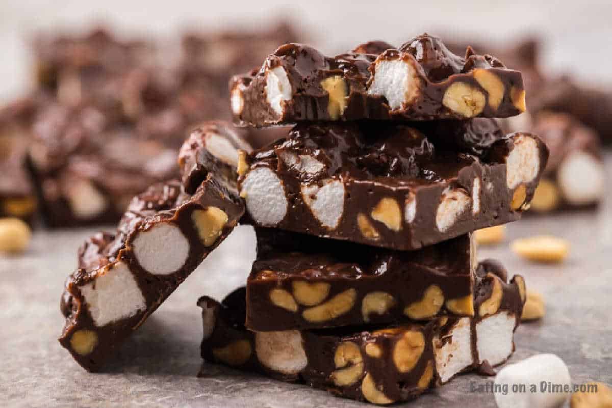 A stack of homemade rocky road candy bars featuring chunks of marshmallows and peanuts, all coated in rich chocolate. In the background, more pieces of this easy rocky road fudge recipe are slightly blurred. The image is set on a textured, neutral-colored surface.