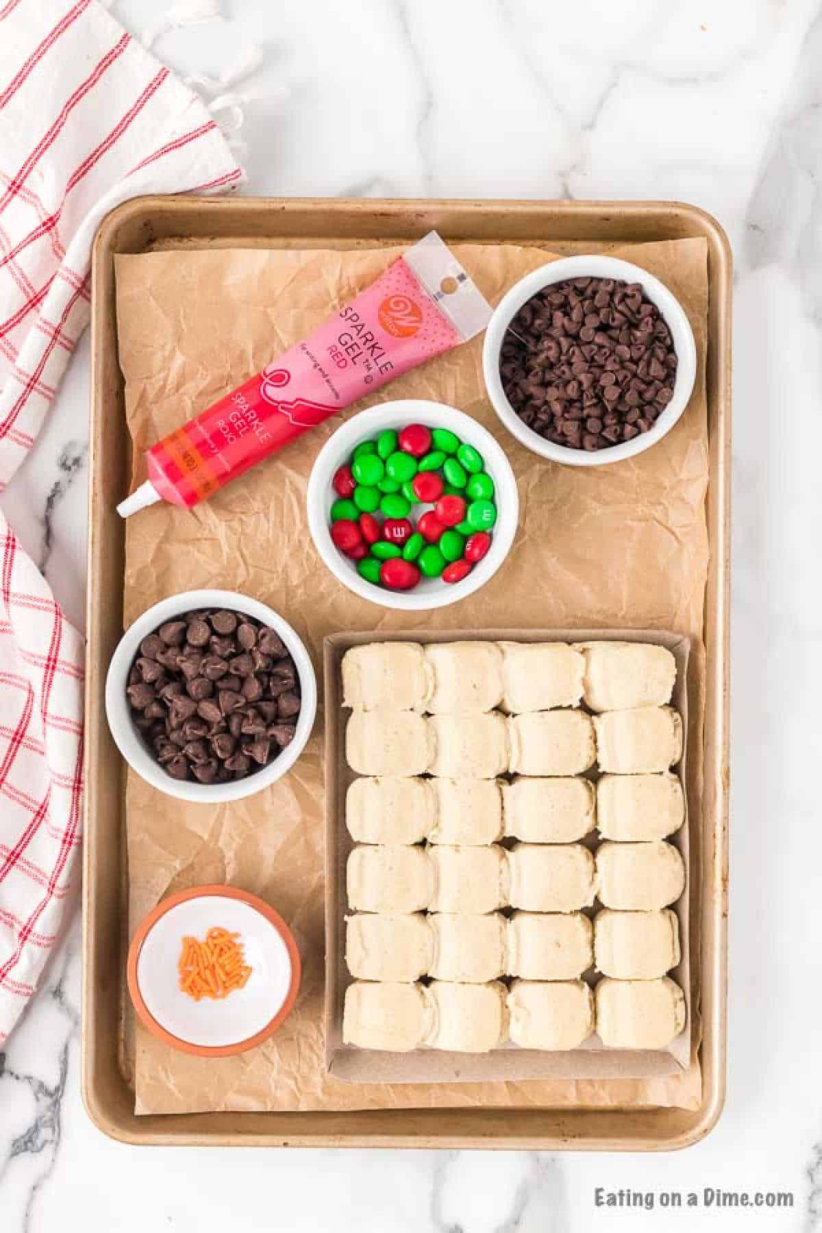 A baking tray lined with parchment paper contains unbaked cookie dough squares, bowls of chocolate chips, and red and green M&M candies for an easy snowman sugar cookie recipe. A tube of red cookie icing and a small dish of orange sprinkles are also on the tray. A red and white cloth is nearby.