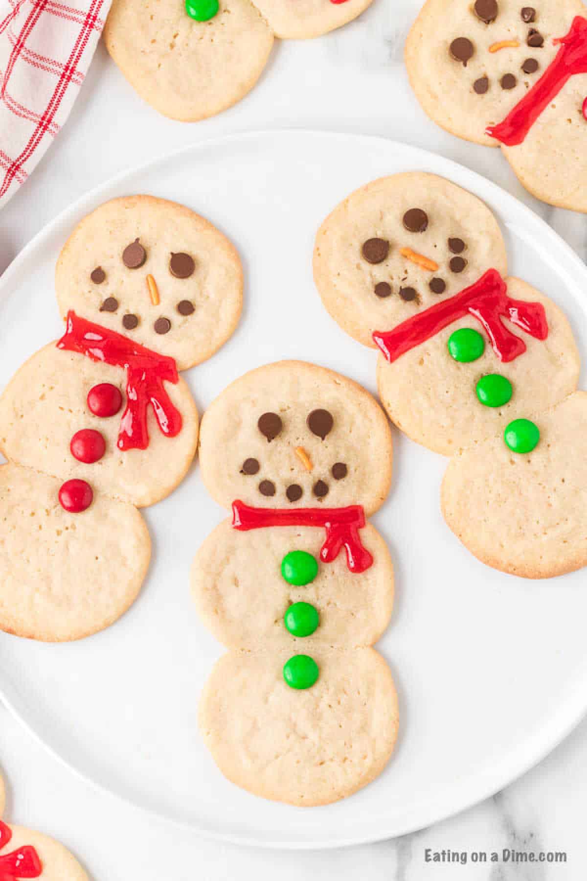 A plate of snowman-shaped cookies, made from an easy snowman sugar cookie recipe, is decorated with red scarves made of icing and buttons made of green and red candies. The snowmen have chocolate chip eyes and mouths, along with an orange candy nose. A red checkered towel is visible in the corner.