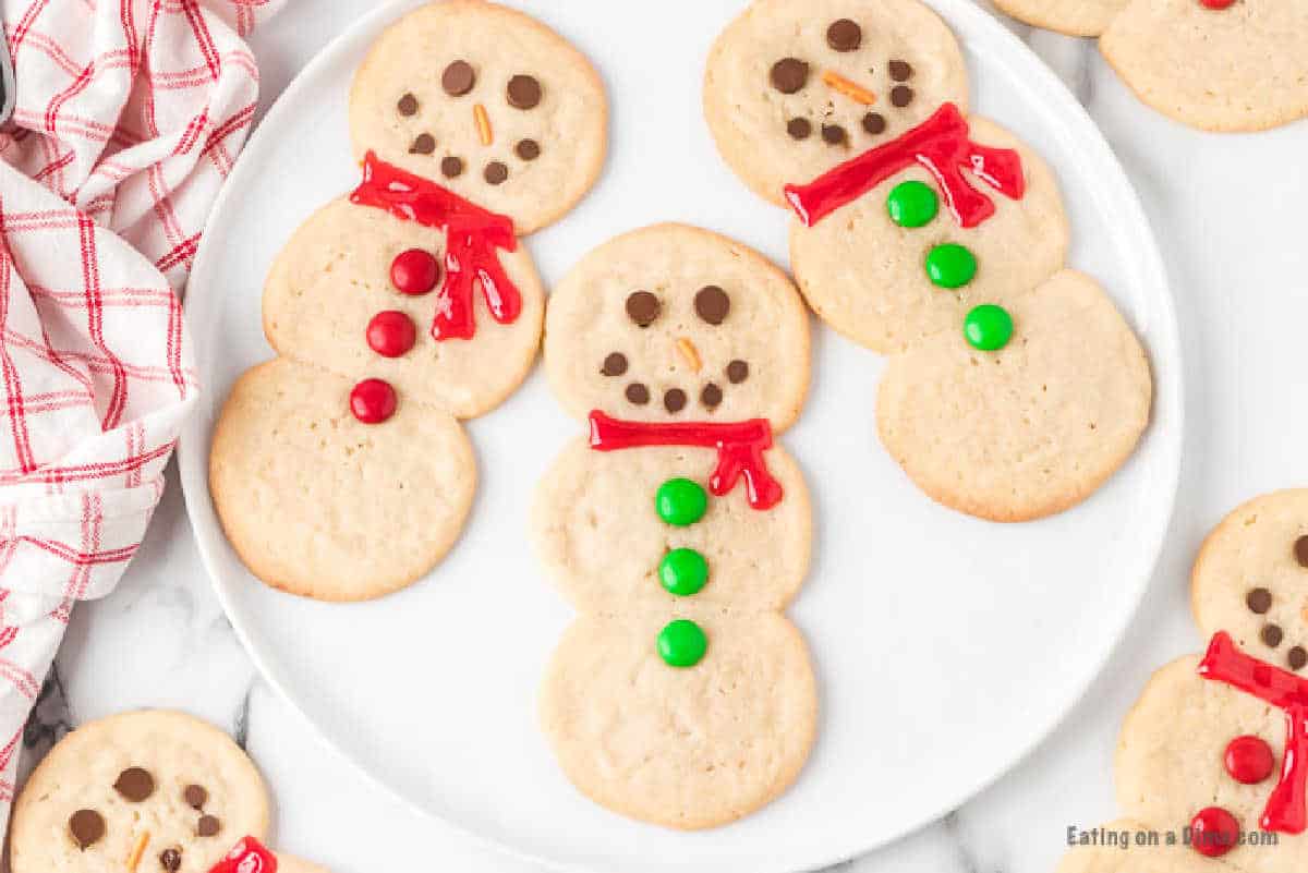 A plate of three snowman-shaped cookies, made using an easy snowman sugar cookie recipe, decorated with chocolate chip eyes and mouths, red or green candy buttons, and red or green icing scarves. Other similar cookies are scattered nearby. A red and white checkered cloth is partially visible on the left.