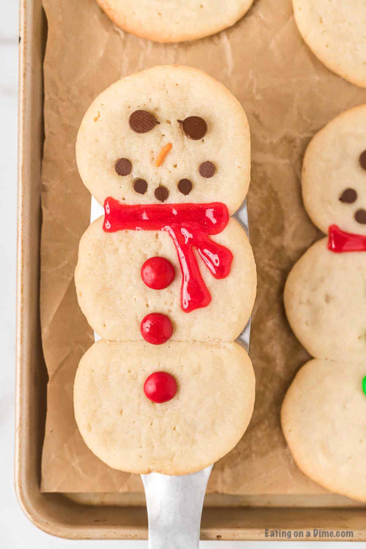 A snowman-shaped cookie is decorated with chocolate chip eyes and mouth, an orange candy for the nose, a red icing scarf, and red button-like candies. The cookie sits on a metal spatula above a parchment-lined baking tray with another similar cookie visible—an easy snowman sugar cookie recipe.