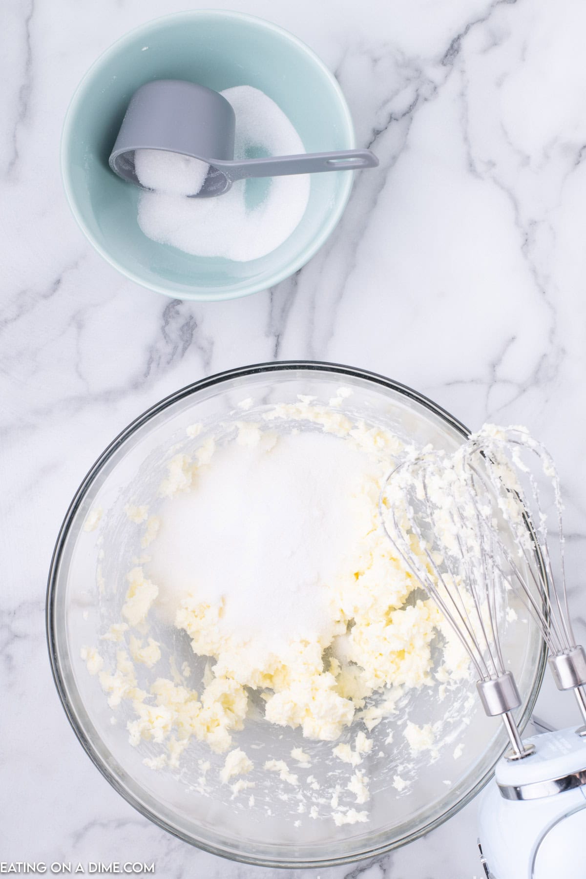 A glass mixing bowl with creamed butter and sugar mixture sits on a marble countertop, ready for the Raspberry Thumbprint Cookie Recipe. An electric hand mixer with batter residue is partially visible on the right. Beside the bowl, there is a small turquoise bowl with sugar and a gray measuring cup.