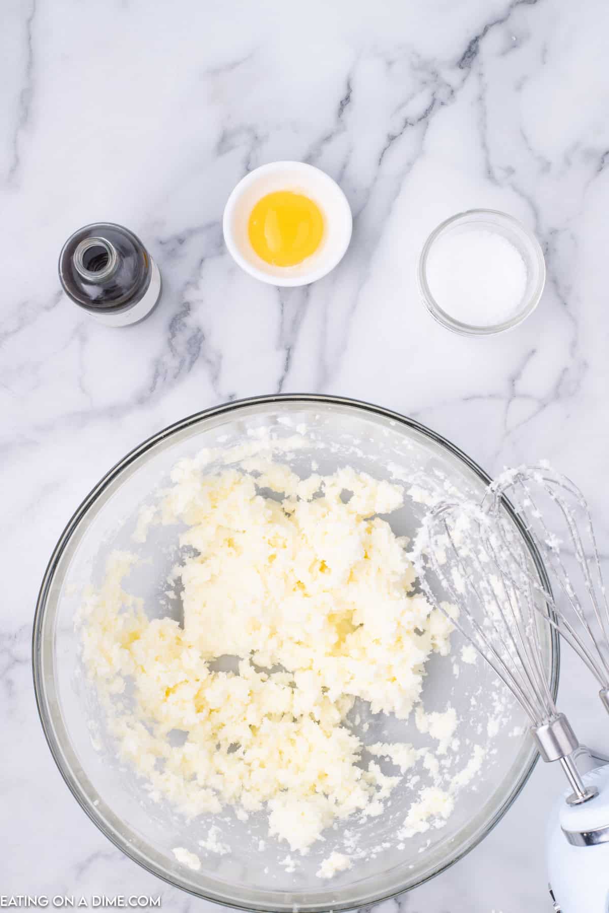 A glass bowl with creamed butter and sugar sits on a marble countertop, next to a whisk, setting the stage for our Raspberry Thumbprint Cookie recipe. Nearby are small containers holding vanilla extract, an egg yolk, and granulated sugar.