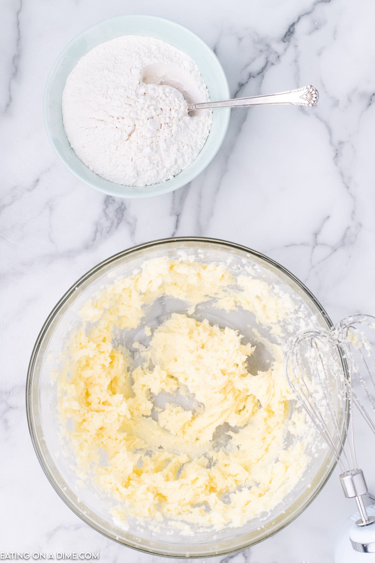 A mixing bowl with creamed butter and sugar sits on a marble countertop, ready for the next step in a Raspberry Thumbprint Cookie recipe. Next to it is a smaller bowl filled with flour and a spoon. A handheld mixer with beaters is partially visible on the right.
