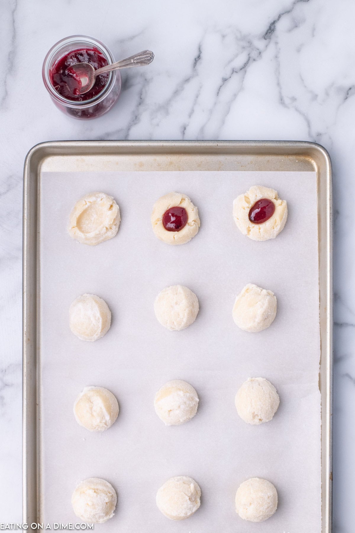 A baking sheet lined with parchment paper holds twelve round cookie dough balls. Two of the cookies have indentations filled with red jam. A jar of red jam with a spoon is placed above the baking sheet on a marble countertop, illustrating our Raspberry Thumbprint Cookie Recipe.