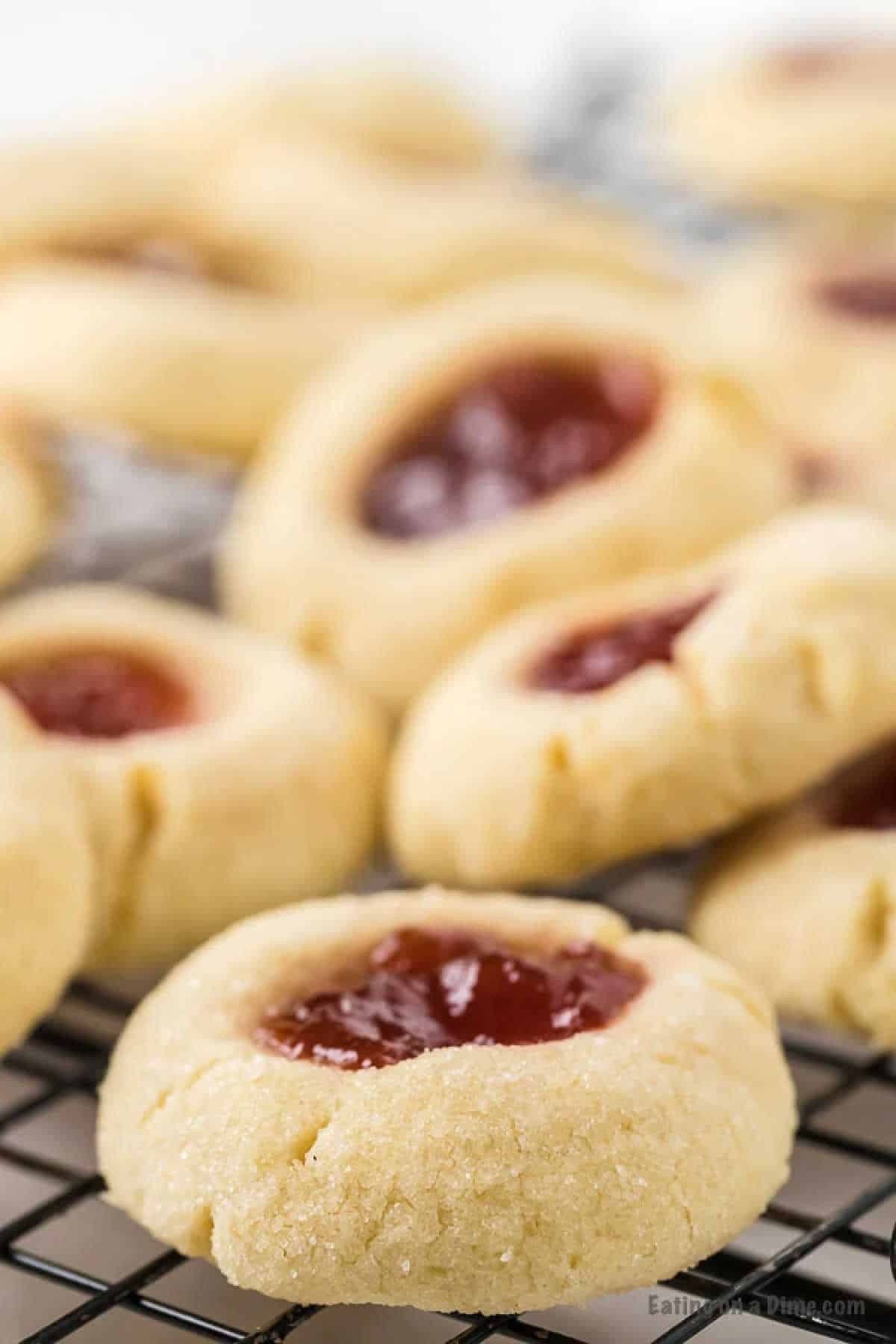 A close-up image of raspberry thumbprint cookies resting on a cooling rack. The cookies have a golden-brown appearance, with a dollop of red jam nestled in the center of each one. The texture appears soft and crumbly, perfect for following your favorite thumbprint cookie recipe.