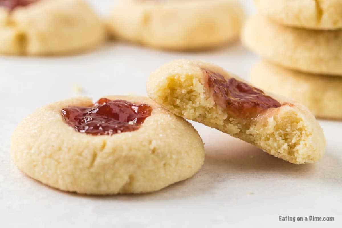 Close-up of thumbprint cookies on a light surface. One cookie is in the foreground, topped with raspberry jam. Beside it, another cookie is broken in half, showing the soft interior. In the background, more cookies and a stack of cookies are slightly out of focus—a perfect Raspberry Thumbprint Cookie Recipe.