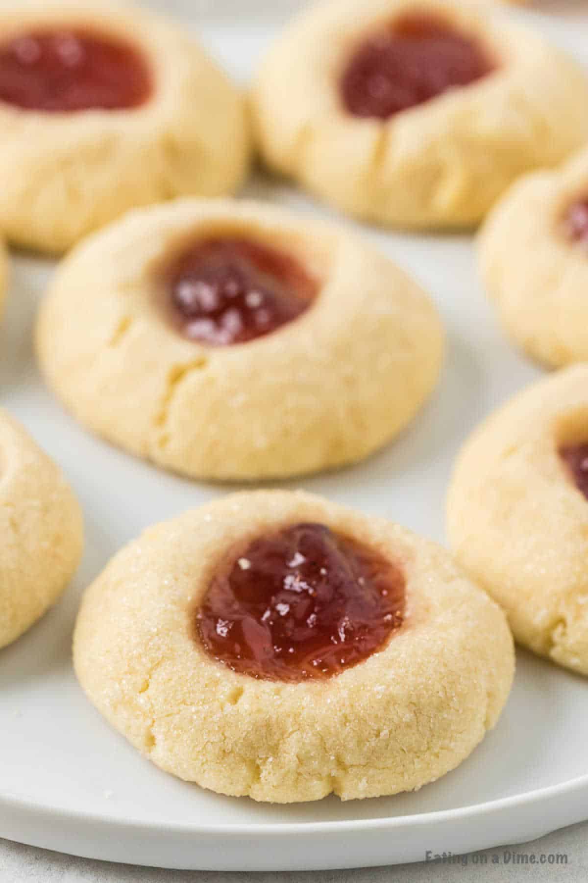 Close-up of several thumbprint cookies on a white plate. The cookies have a golden-brown, crumbly appearance with a dollop of red jam in the center of each, giving them a soft, homemade look—just like from a Raspberry Thumbprint Cookie Recipe.